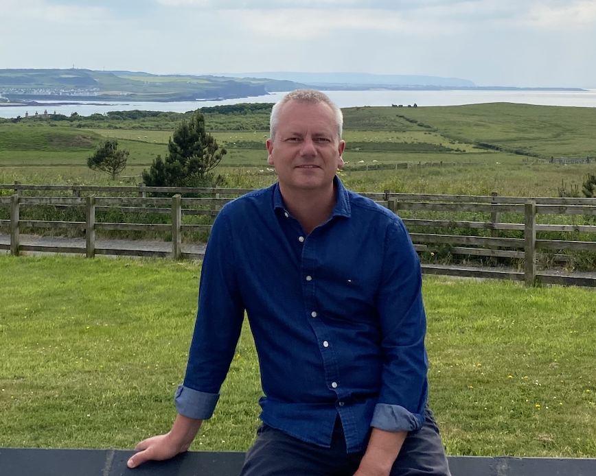 A man in a blue shirt sitting on a black railing outdoors with a green field, trees, and a river in the background on a cloudy day.