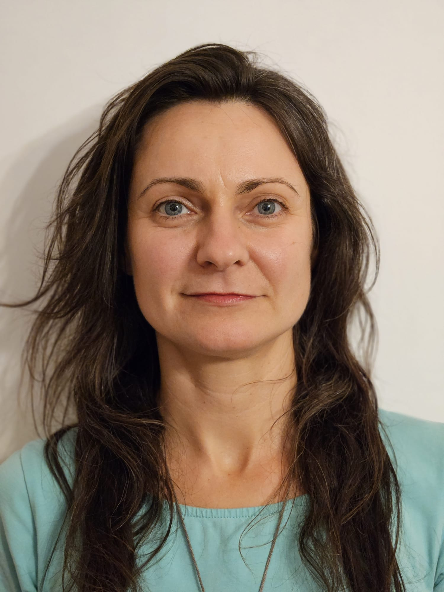 A woman with long brown hair, blue eyes, and a light blue shirt standing against a plain white wall.