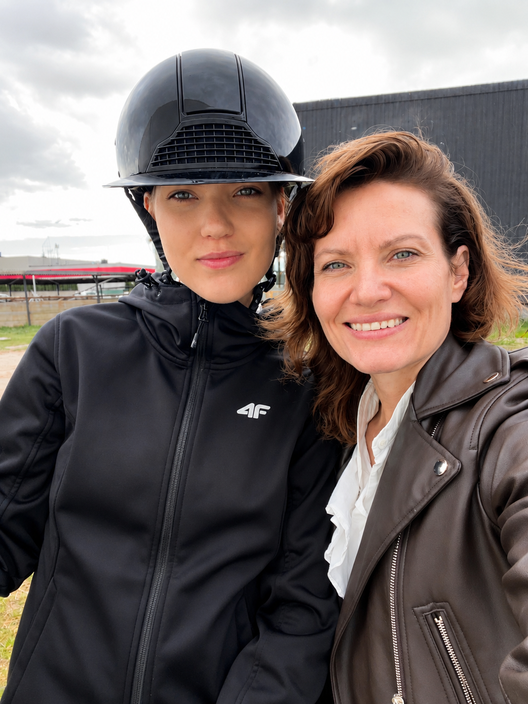 Two women taking a selfie outdoors on a cloudy day, with one wearing a motorcycle helmet and jacket, and the other in a leather jacket.