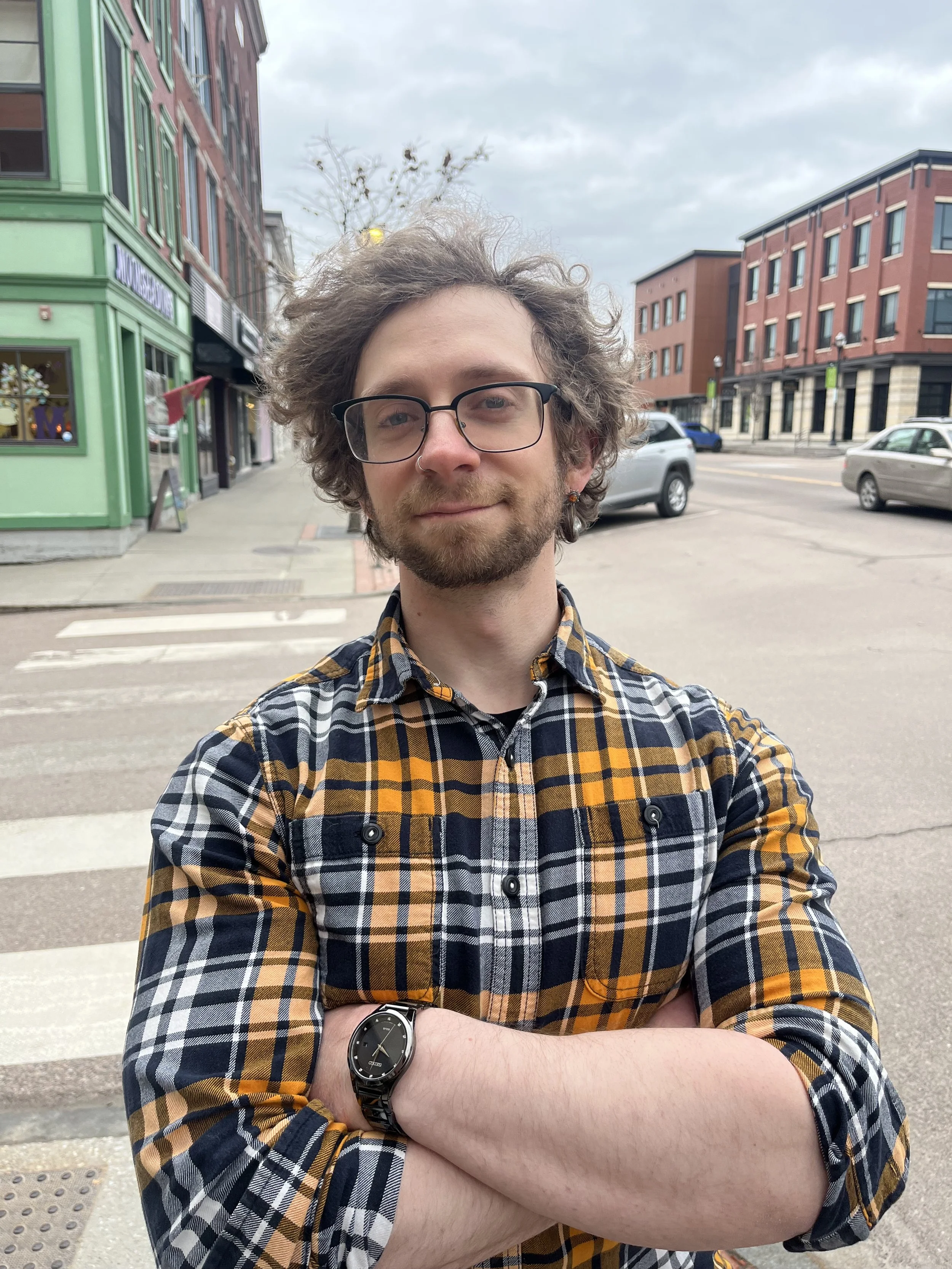 A man with curly hair, glasses, and a beard standing on a city street with his arms crossed, wearing a plaid shirt and a watch, during daytime with overcast sky.