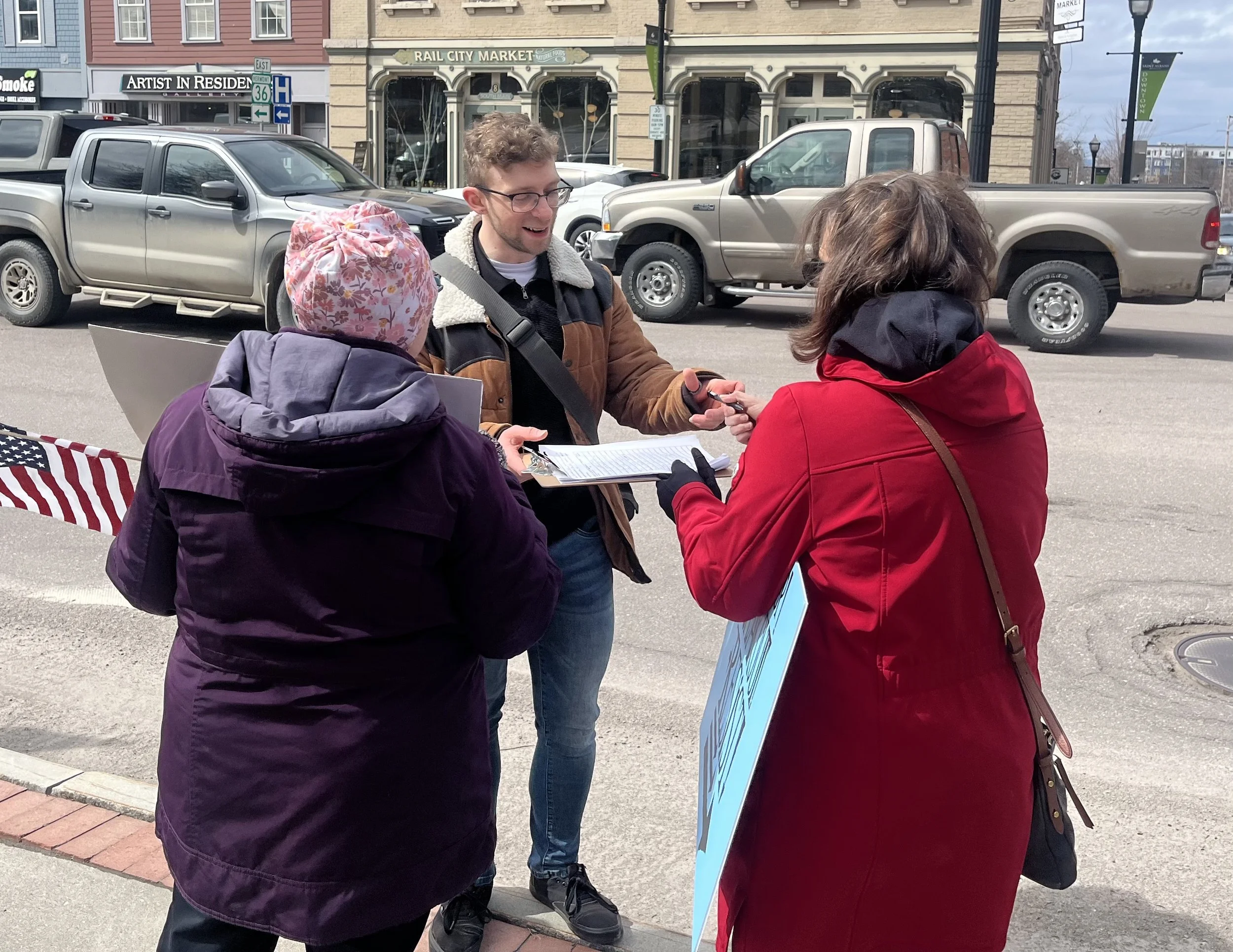 A man is speaking to two women on a sidewalk near a street with parked cars. One woman is wearing a purple jacket and pink floral hat, holding a clipboard. The other woman, in a red jacket, is holding a blue sign. The background shows storefronts and parked vehicles.