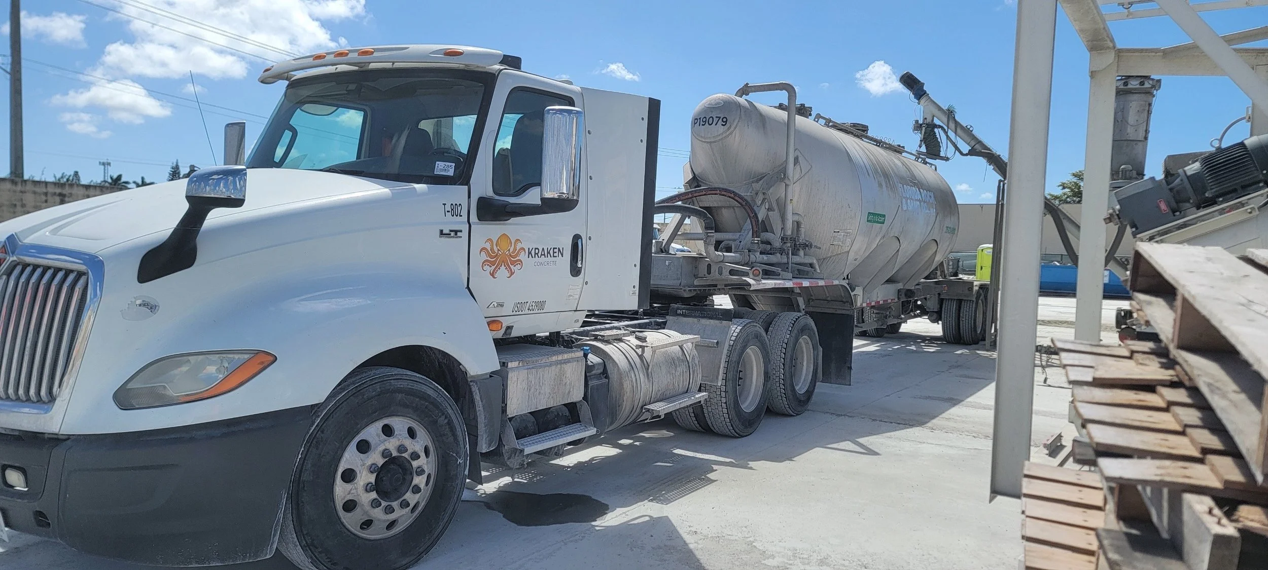 White cement mixer truck parked outdoors under a blue sky with clouds; the truck has a logo reading 'Kraken Concrete' on the door, and construction materials are visible on the right side.