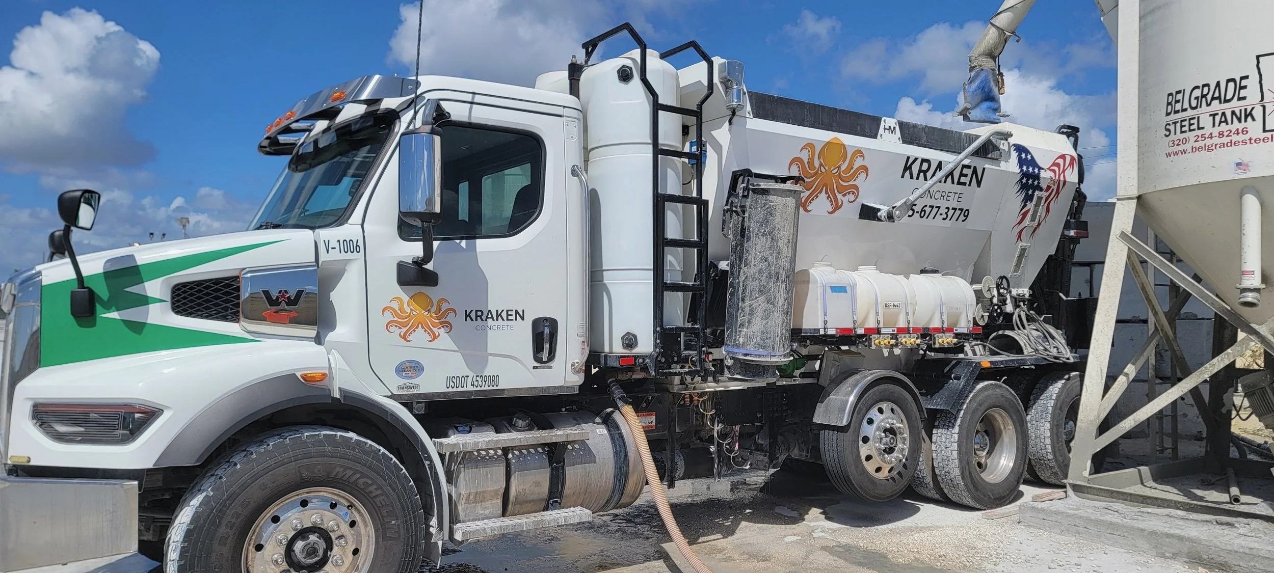 Concrete mixer truck with company logos and equipment at a construction site under a partly cloudy sky.