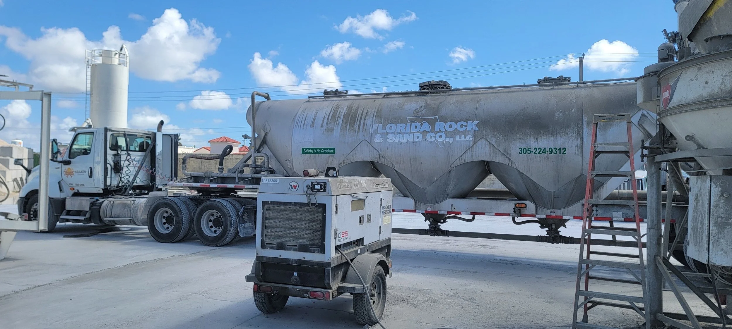 A cement or sand delivery truck with a silver tank labeled 'Florida Rock & Sand Co. LLC' parked on a concrete lot, with a ladder on the side, under a blue sky with clouds.