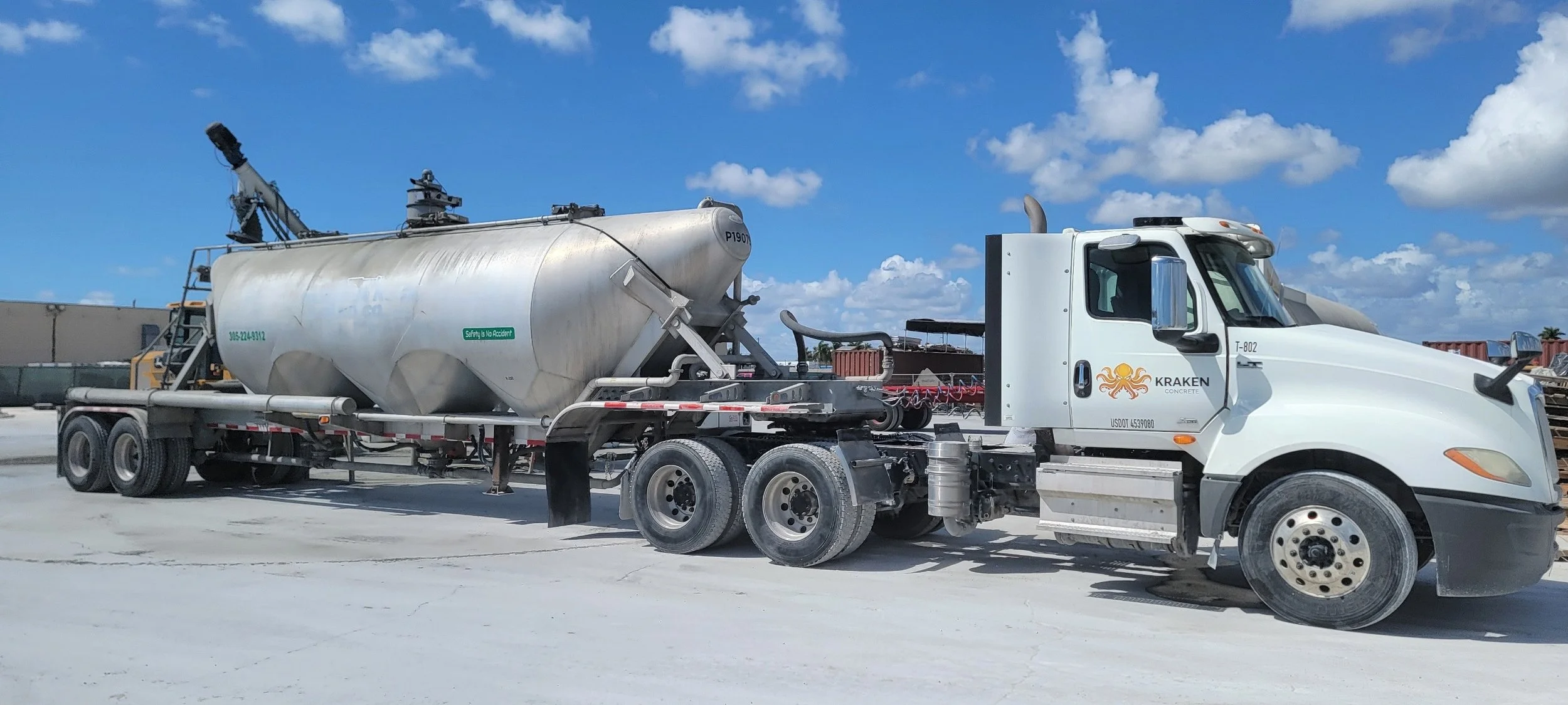 White concrete mixer truck with tank, labeled 'Kraken Concrete' and a crane, parked on a construction site under a blue sky with clouds.