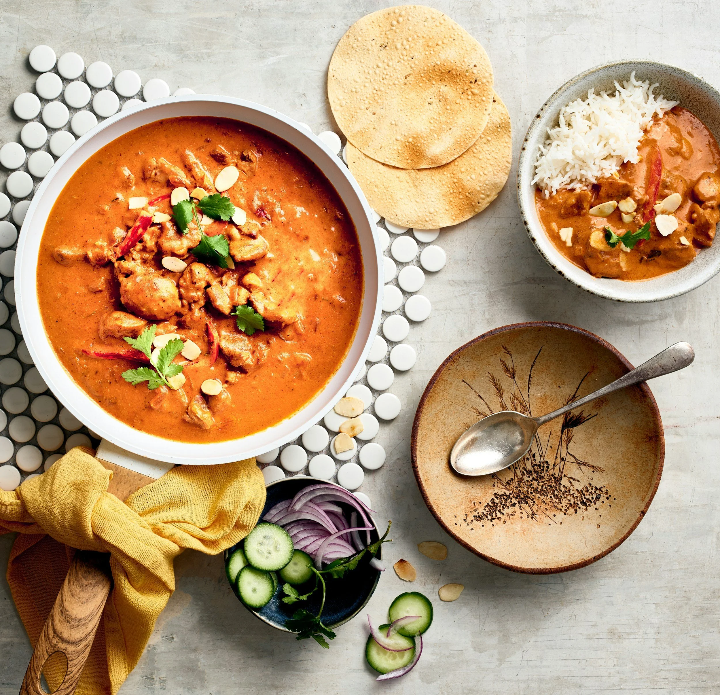 A table with various ingredients and dishes, including a bowl of chicken curry garnished with cilantro and almonds, a bowl of rice with curry, two papadums, sliced cucumber, onion, a yellow cloth, and a spoon with black peppercorns.