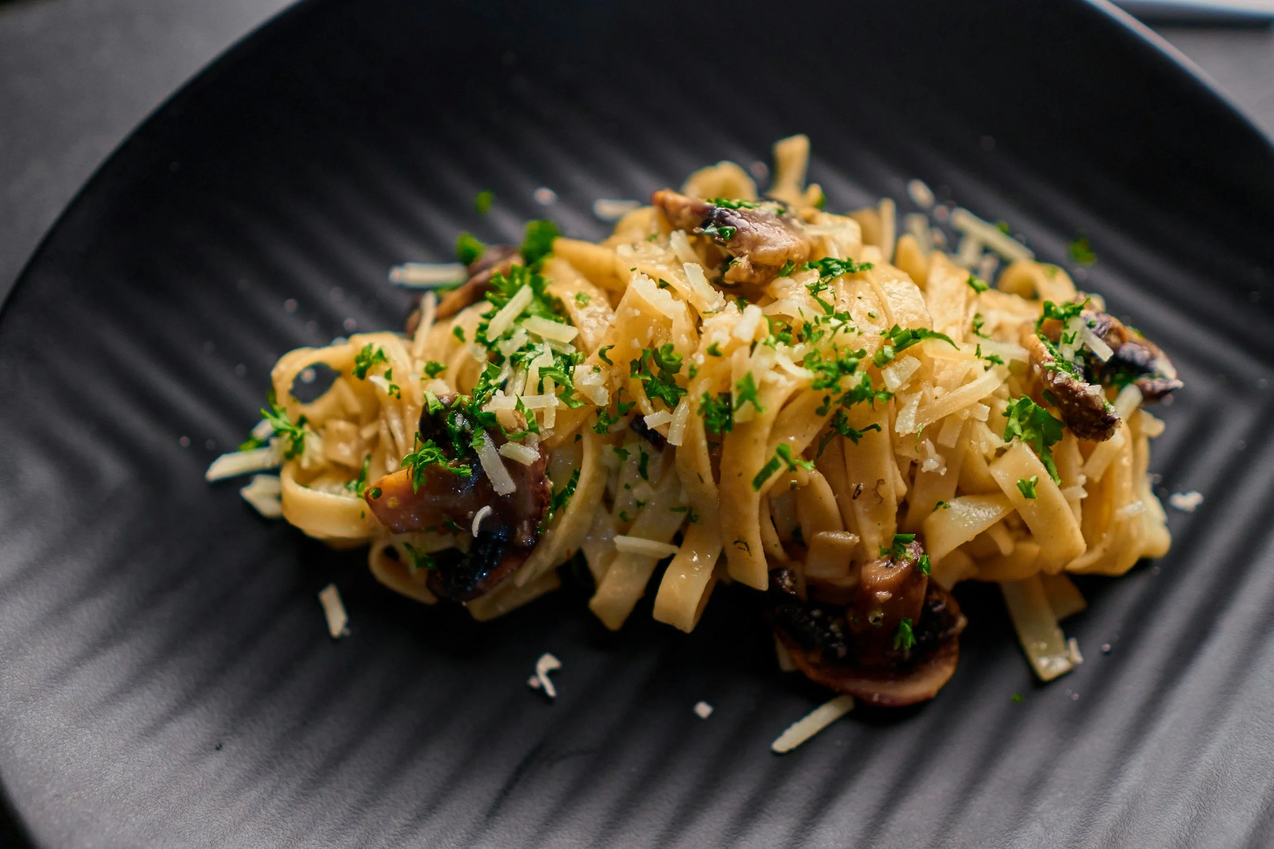 Close-up of a plate of fettuccine pasta with mushrooms and grated cheese, garnished with chopped parsley on a black textured surface.