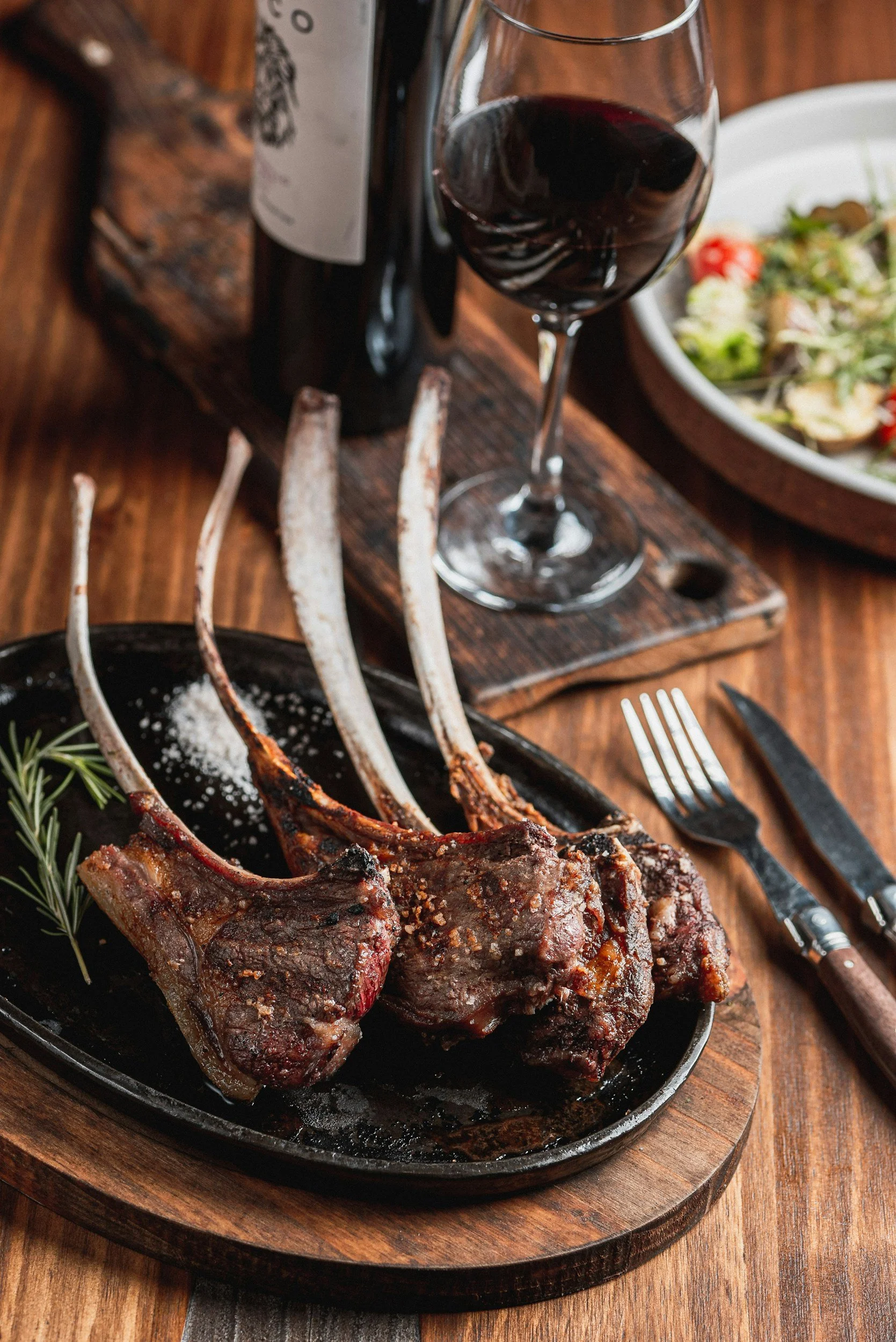 Close-up of cooked lamb chops with a rosemary garnish on a black plate, placed on a wooden table.