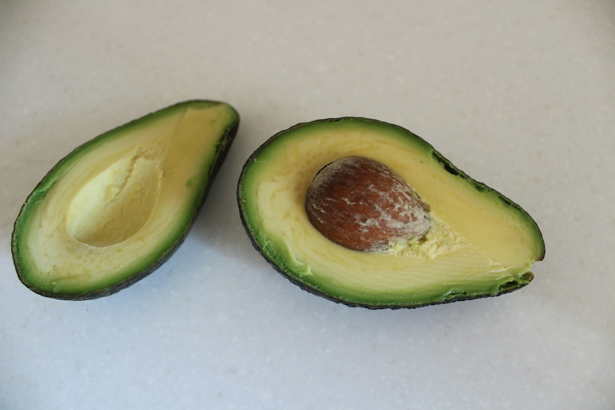 A halved avocado with green outer skin, yellow-green flesh, and a large brown seed inside, placed on a white surface.