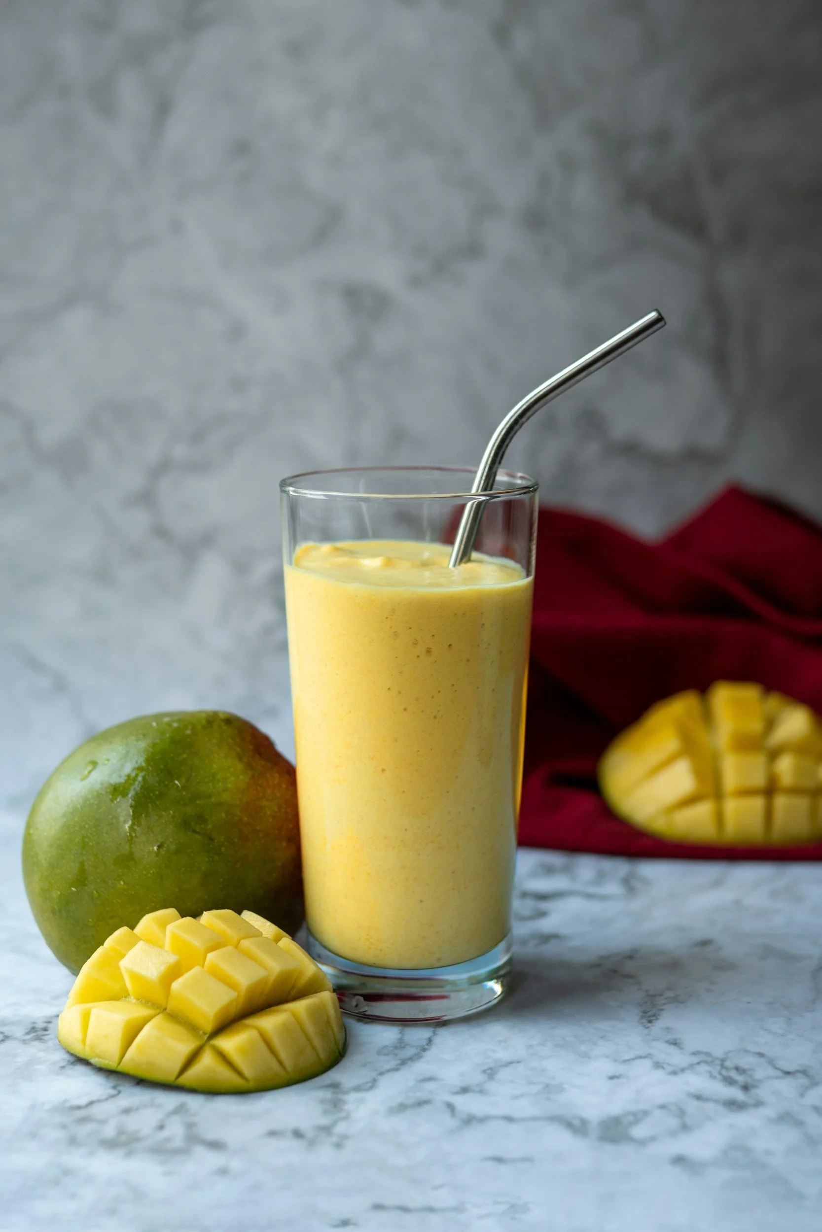 A glass of mango smoothie with a metal straw on a marble surface, with a whole mango, mango chunks, and a sliced mango in the background, and a red cloth in the background.