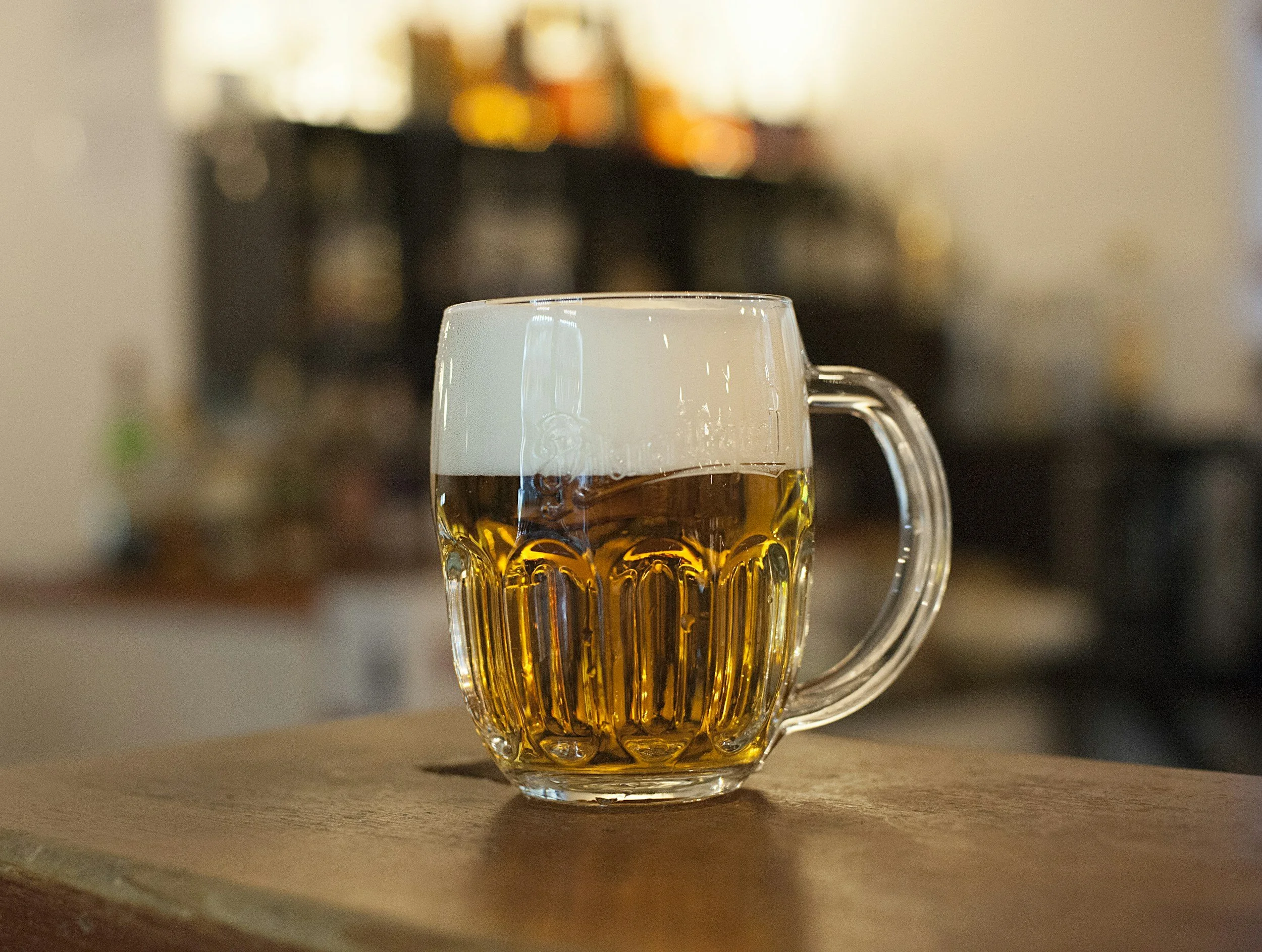 A clear glass mug filled with beer topped with foam, placed on a wooden surface with a blurred background of bottles.