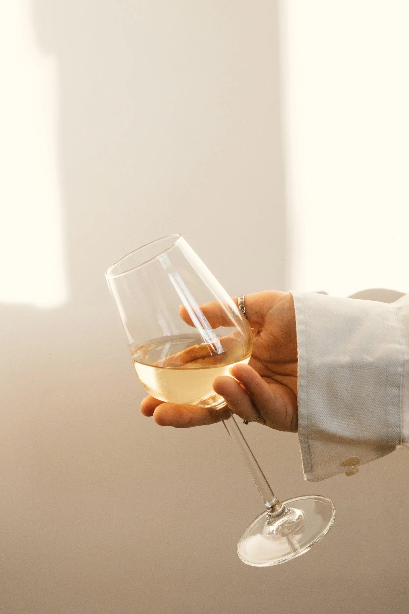 A person is holding a glass of white wine, with their hand partially visible, wearing a white shirt sleeve. The background is plain and light-colored.