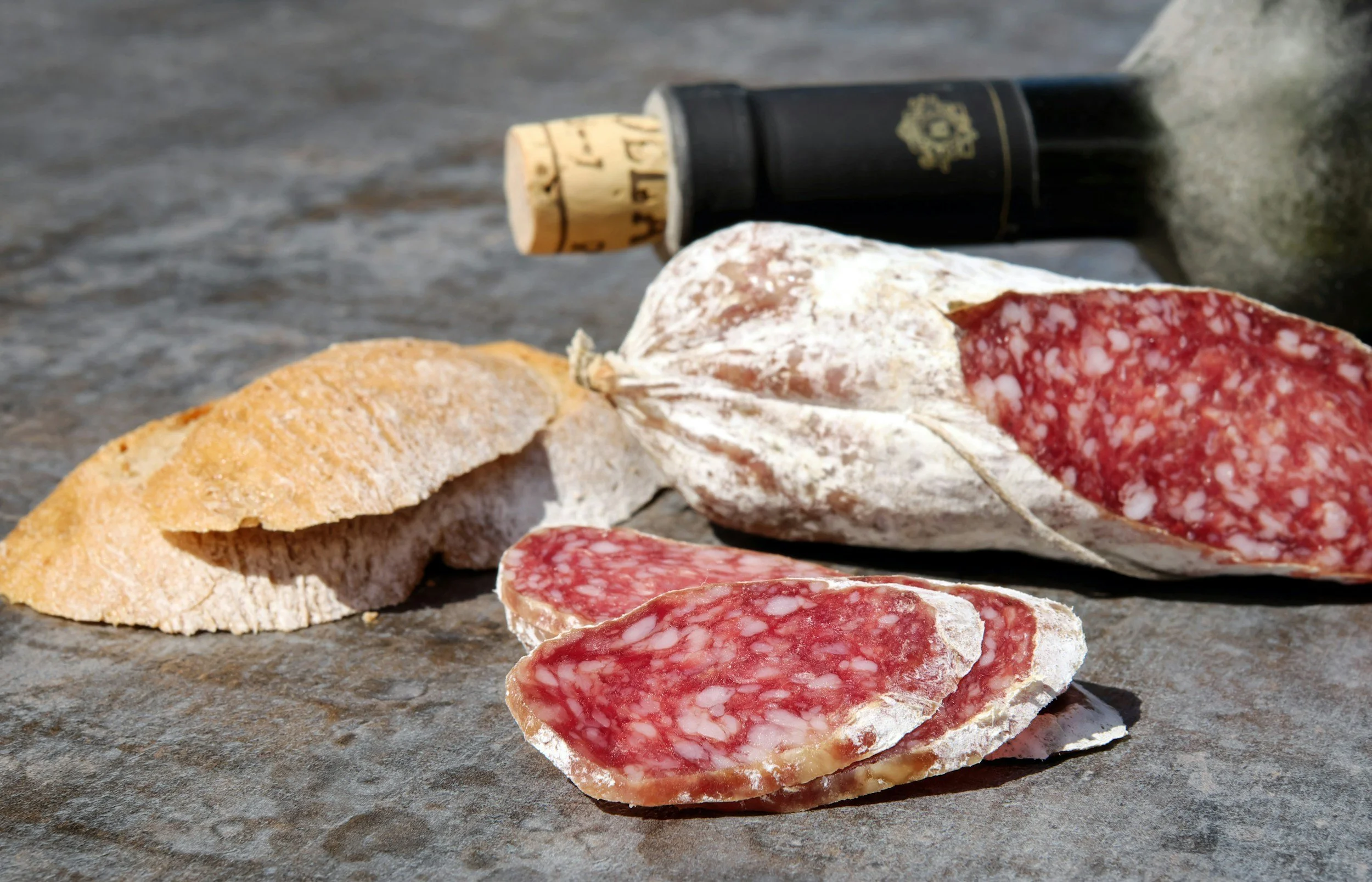 Slices of dry-cured salami and a piece of crusty bread on a stone surface, with a small bottle of dark-colored liquid in the background.