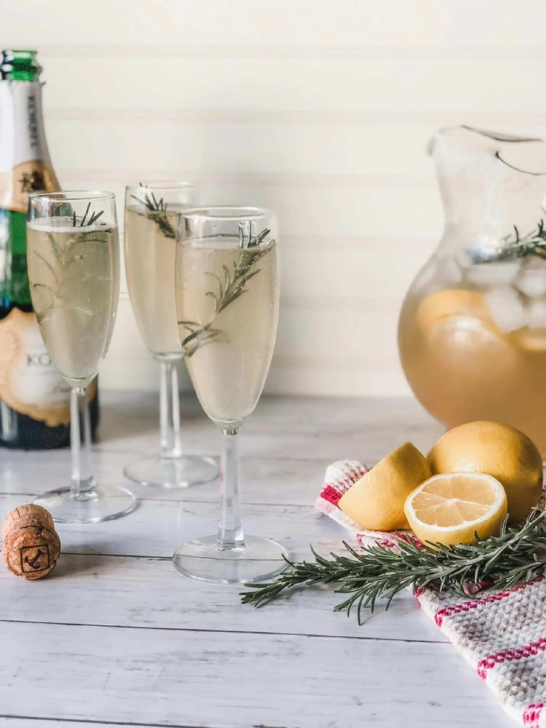 Three champagne flutes with sprigs of rosemary and bubbly liquid, a sliced lemon, a wine carafe with more rosemary and lemon, a cork, and a partially visible bottle in the background, all set on a white wooden surface with a red and white cloth.