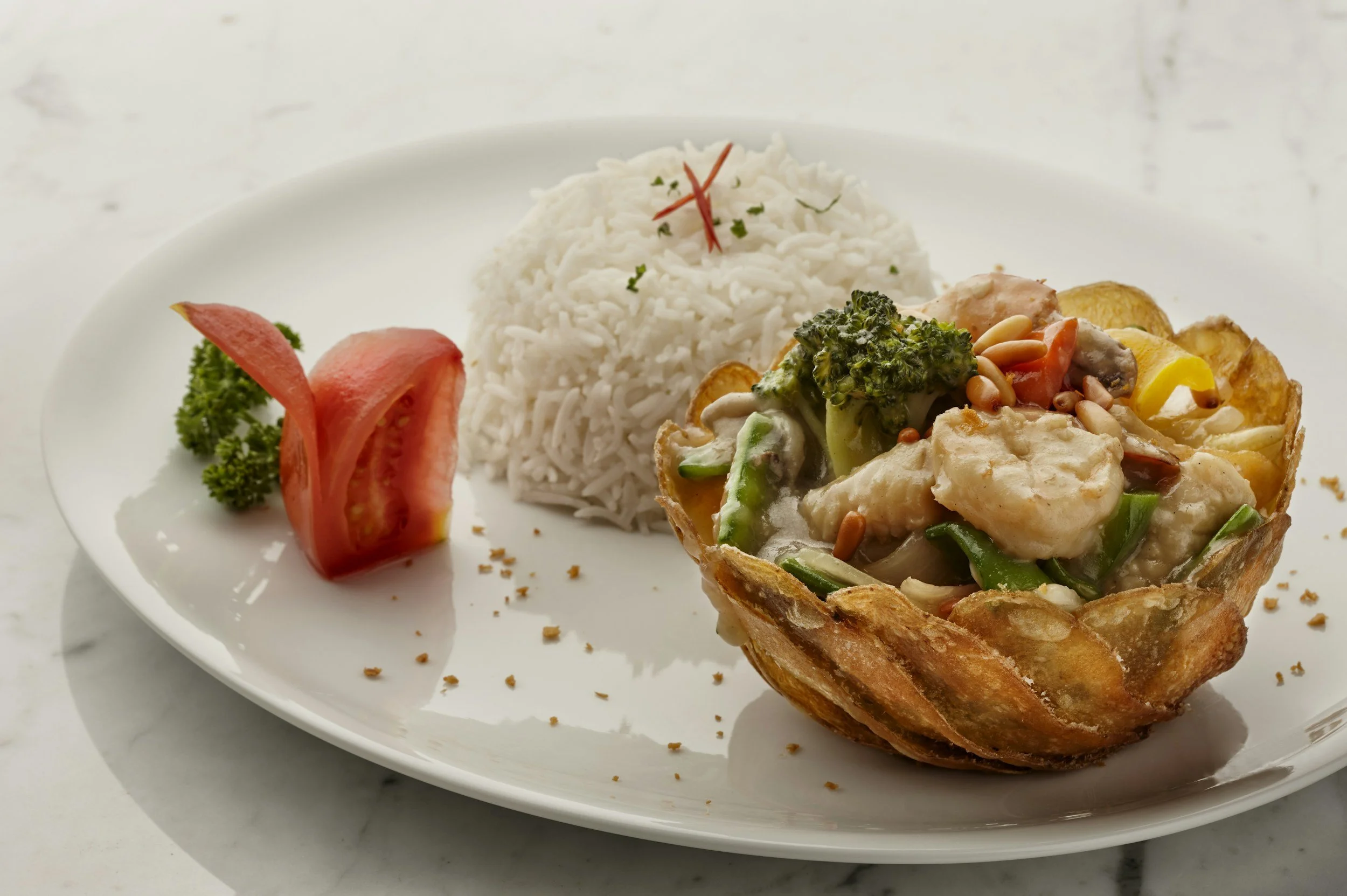 A white plate with a serving of rice and a vegetable and chicken dish in a crispy edible bowl, garnished with sprigs of parsley and a sliced tomato with lettuce.