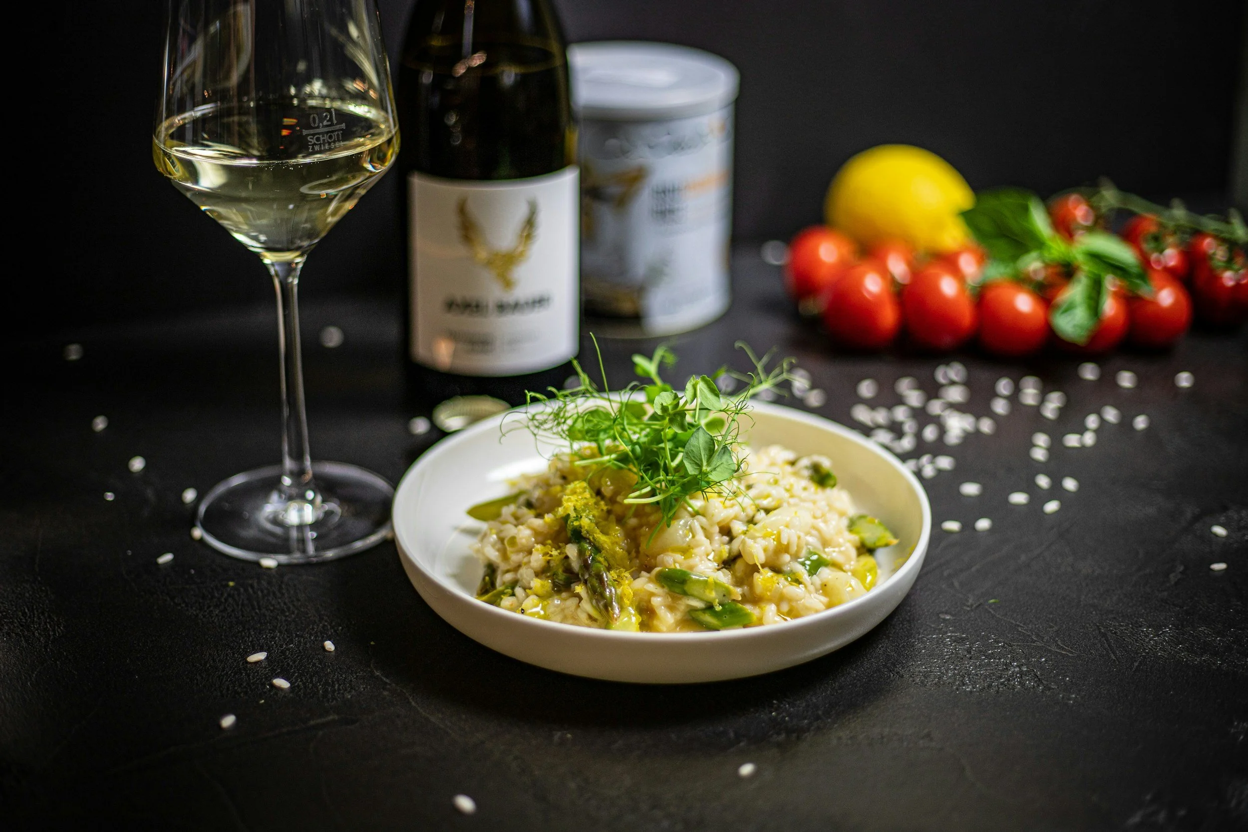 A glass of white wine, a bottle of white wine, canned food, a bunch of cherry tomatoes, a lemon, and a bowl of pasta garnished with green herbs, all on a black surface with scattered rice grains.