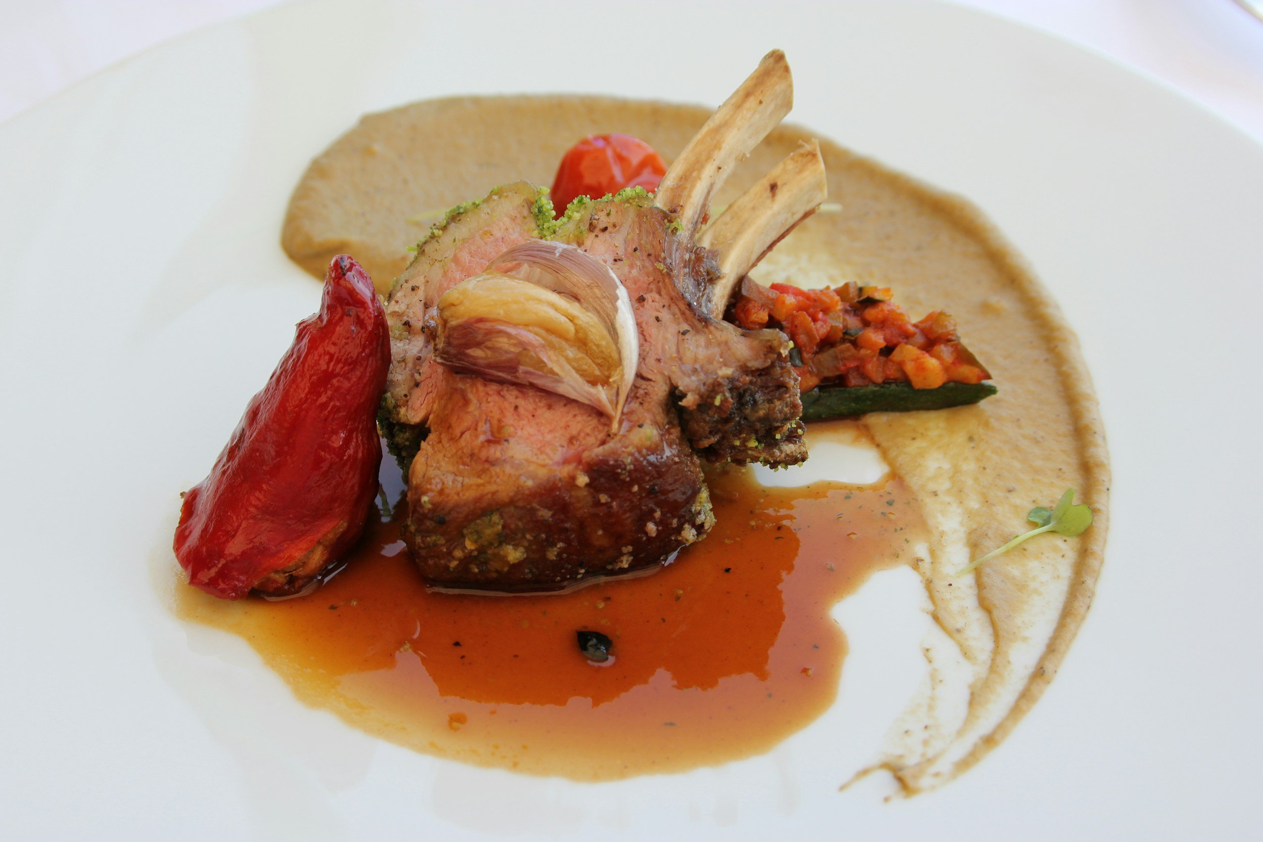 A plated gourmet dish featuring a rack of lamb with sauce, roasted red peppers, and a dollop of sauce with herbs on a white plate.