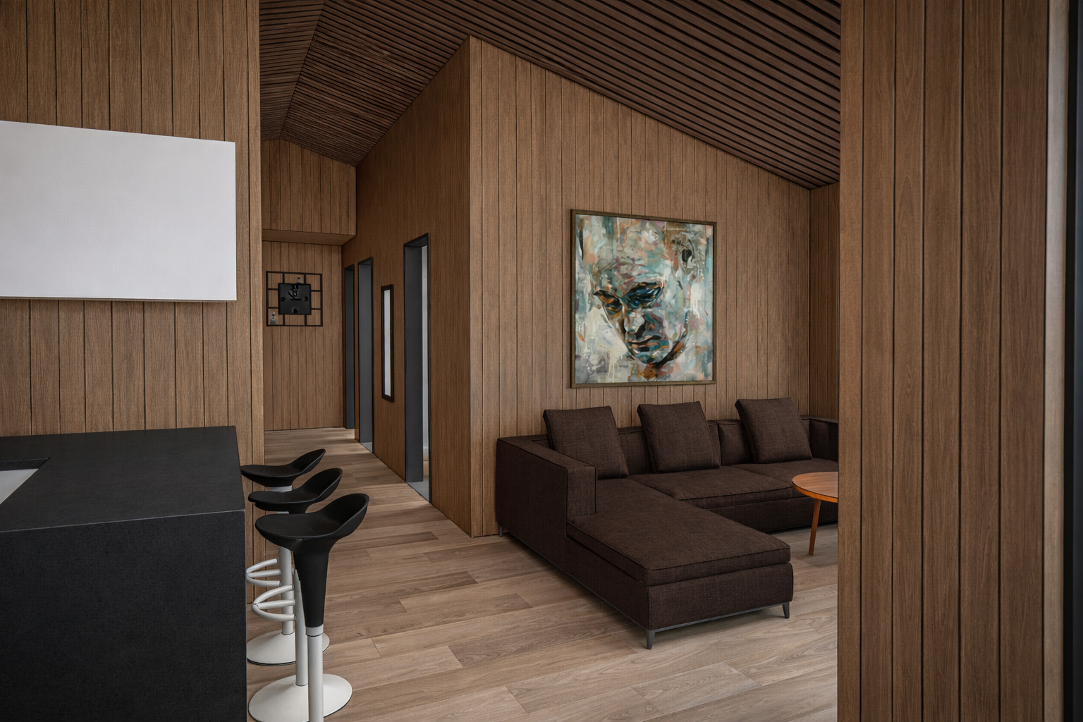 Modern interior with wood-paneled walls and ceiling, featuring a dark brown sectional sofa with cushions, a framed colorful abstract portrait painting on the wall, a round wooden coffee table, and black bar stools with white bases at a black counter, all on light wood flooring.