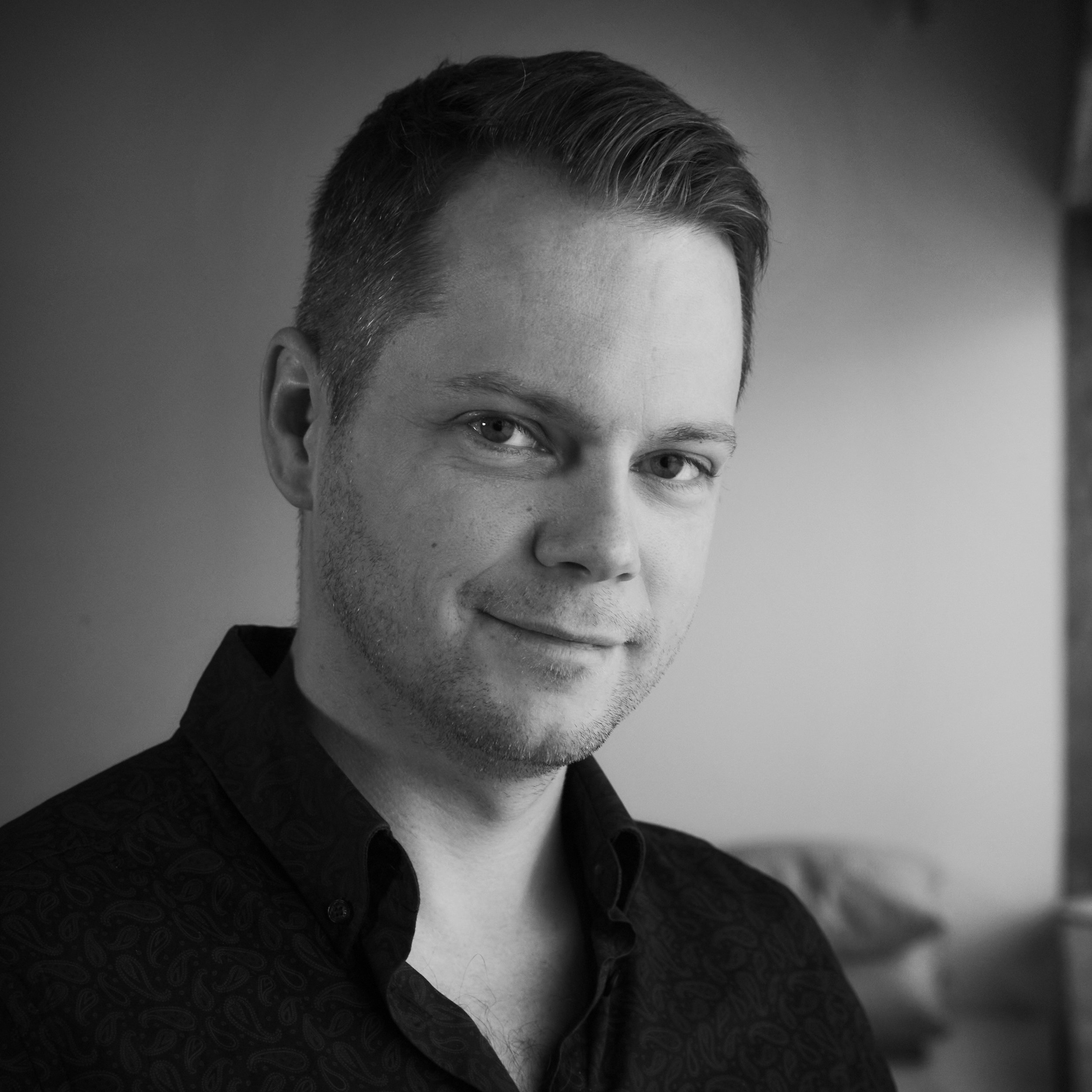 Black and white close-up portrait of a man with short, styled hair, smiling slightly, wearing a dark patterned shirt.
