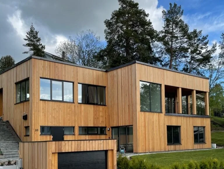 Modern two-story house with wooden siding, large windows, and a balcony, situated in a green landscape surrounded by tall trees.