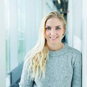Portrait of a young woman with long blonde hair wearing a gray sweater, smiling in a modern indoor setting.