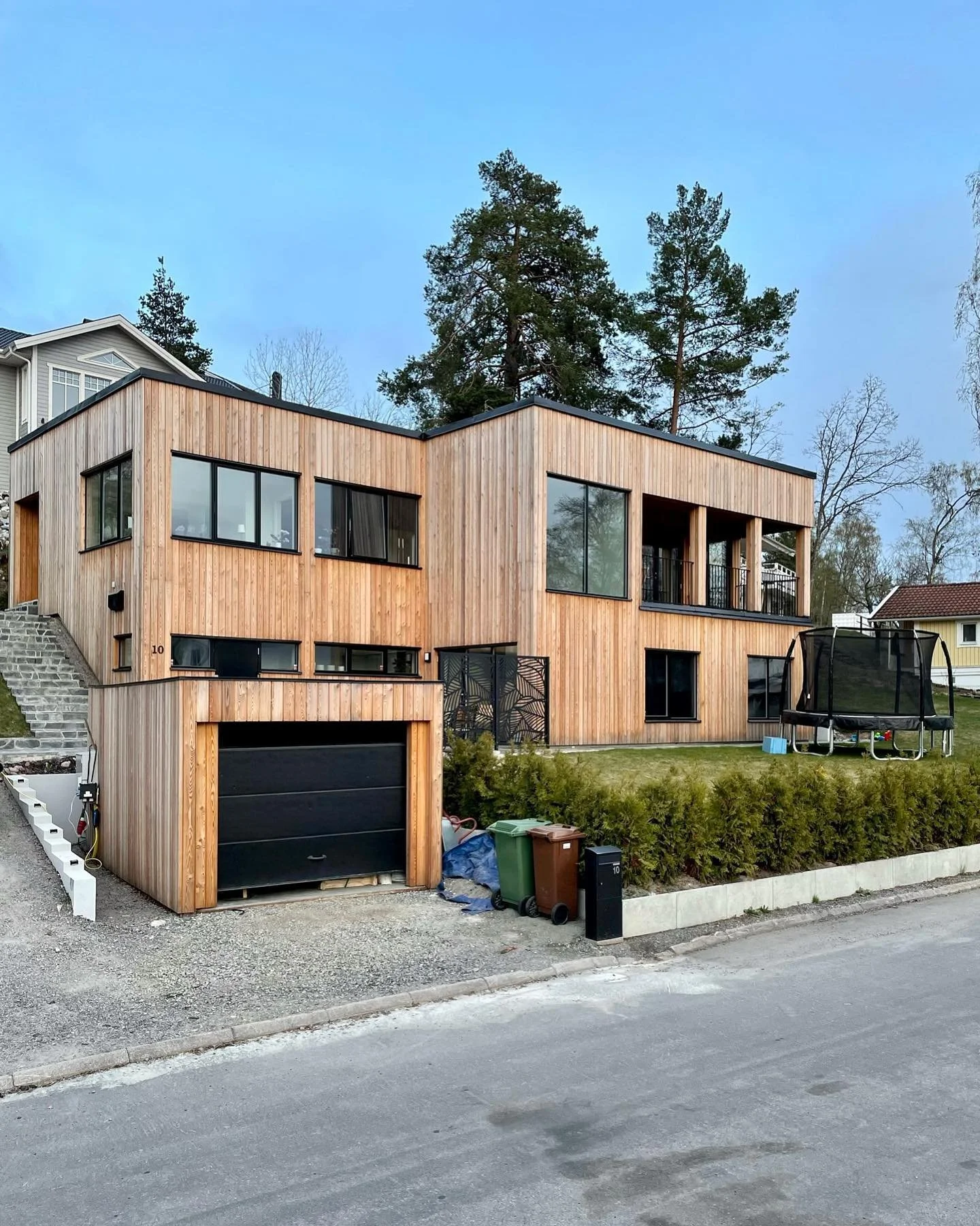 Modern two-story house with wooden exterior, large windows, and a balcony. Front yard has bushes, trash bins, and a trampoline, with a driveway and neighboring houses visible.