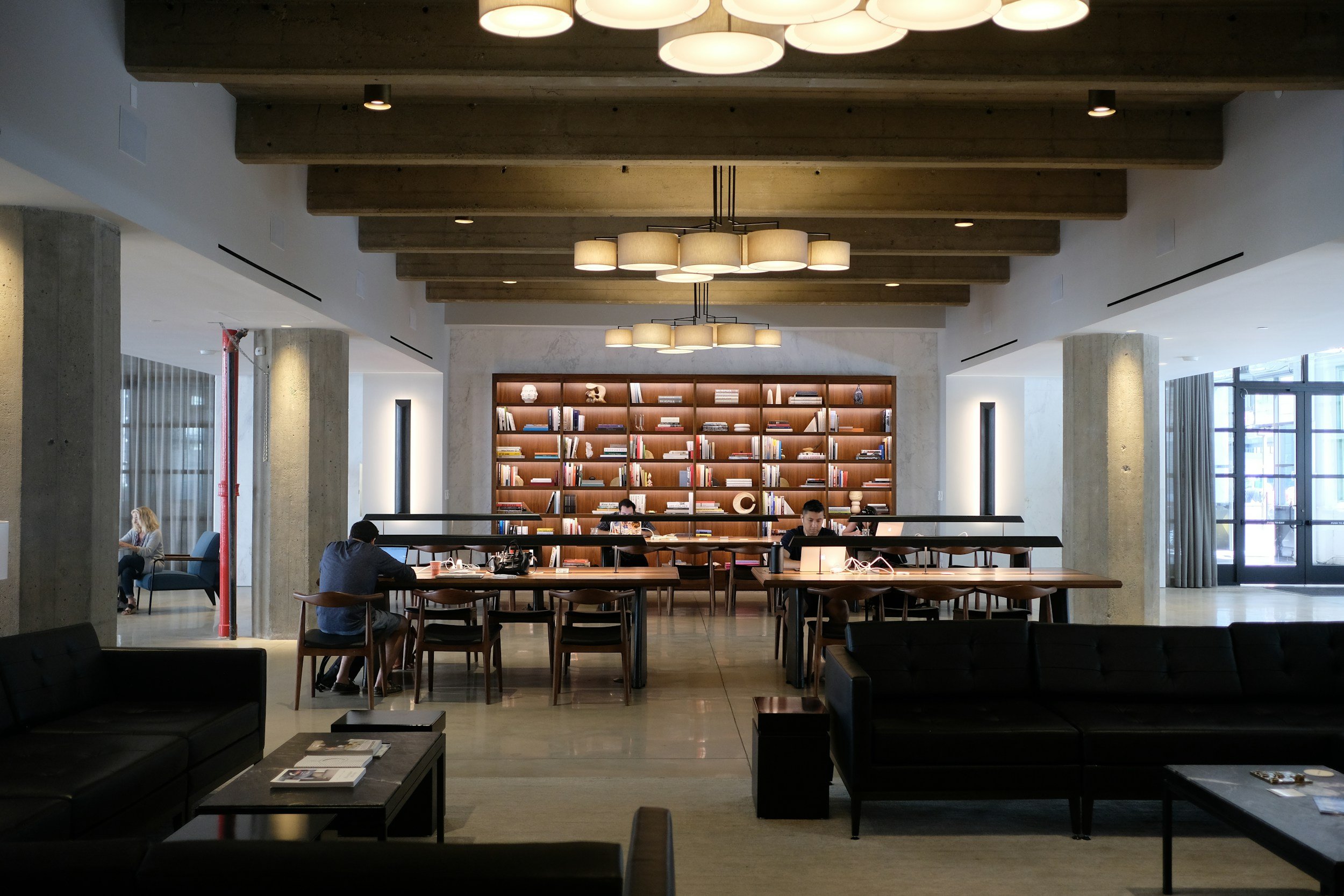 A spacious modern library or coworking space with high ceilings and exposed wooden beams. There is a large illuminated ceiling fixture, a tall bookshelf filled with books, and several people working at desks with laptops. Windows with curtains let in natural light.