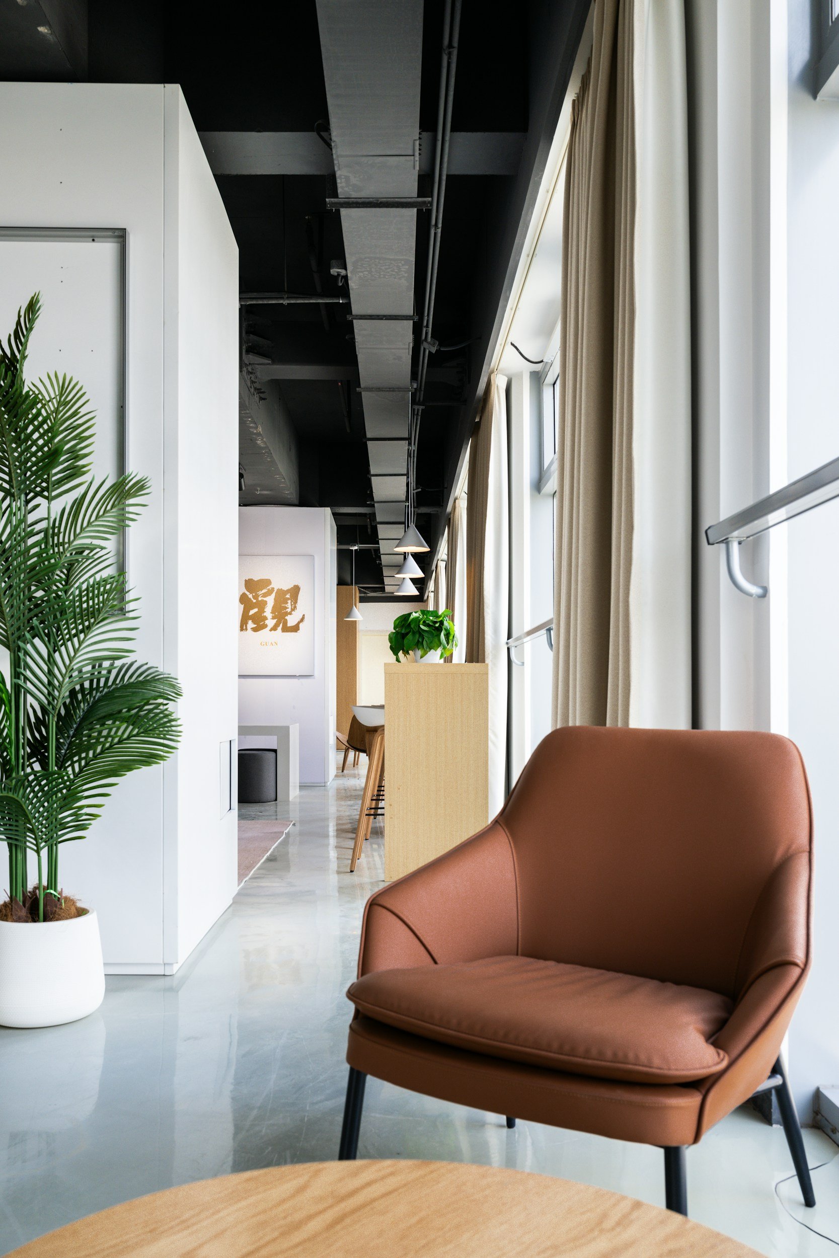 A modern, cozy cafe or lounge area with natural light, beige curtains, green plants, a brown comfortable armchair, and wooden tables.
