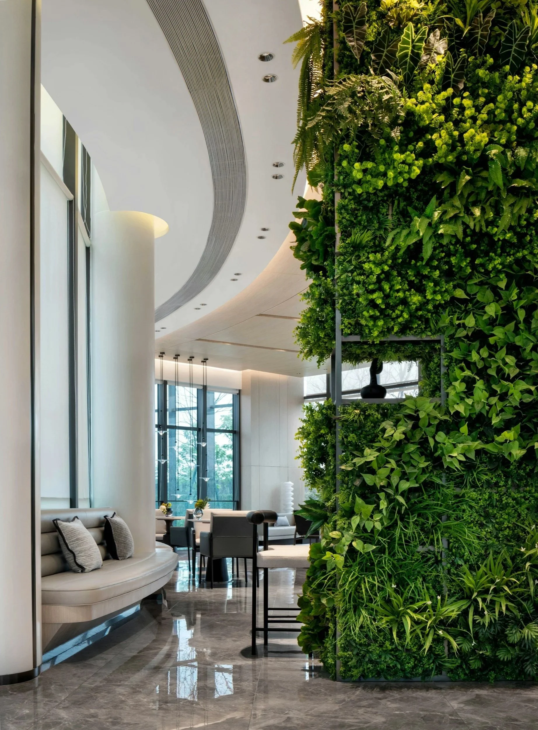 Modern interior lobby or lounge area with a green wall of lush plants, white curved seating with pillows, and large windows allowing natural light to fill the space.