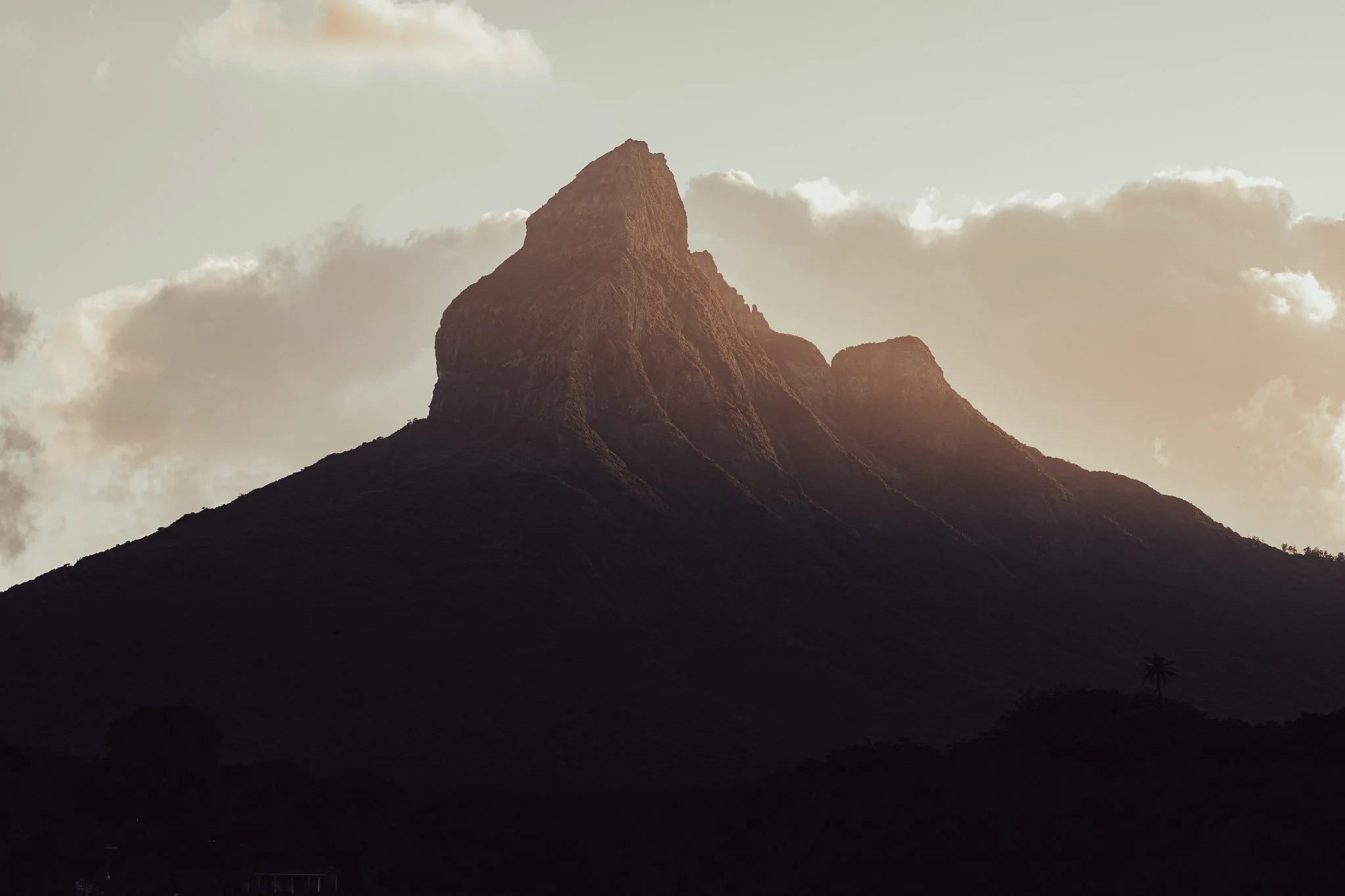 A large mountain with a steep pointed peak, surrounded by clouds, with a dark foreground and small trees at the base.