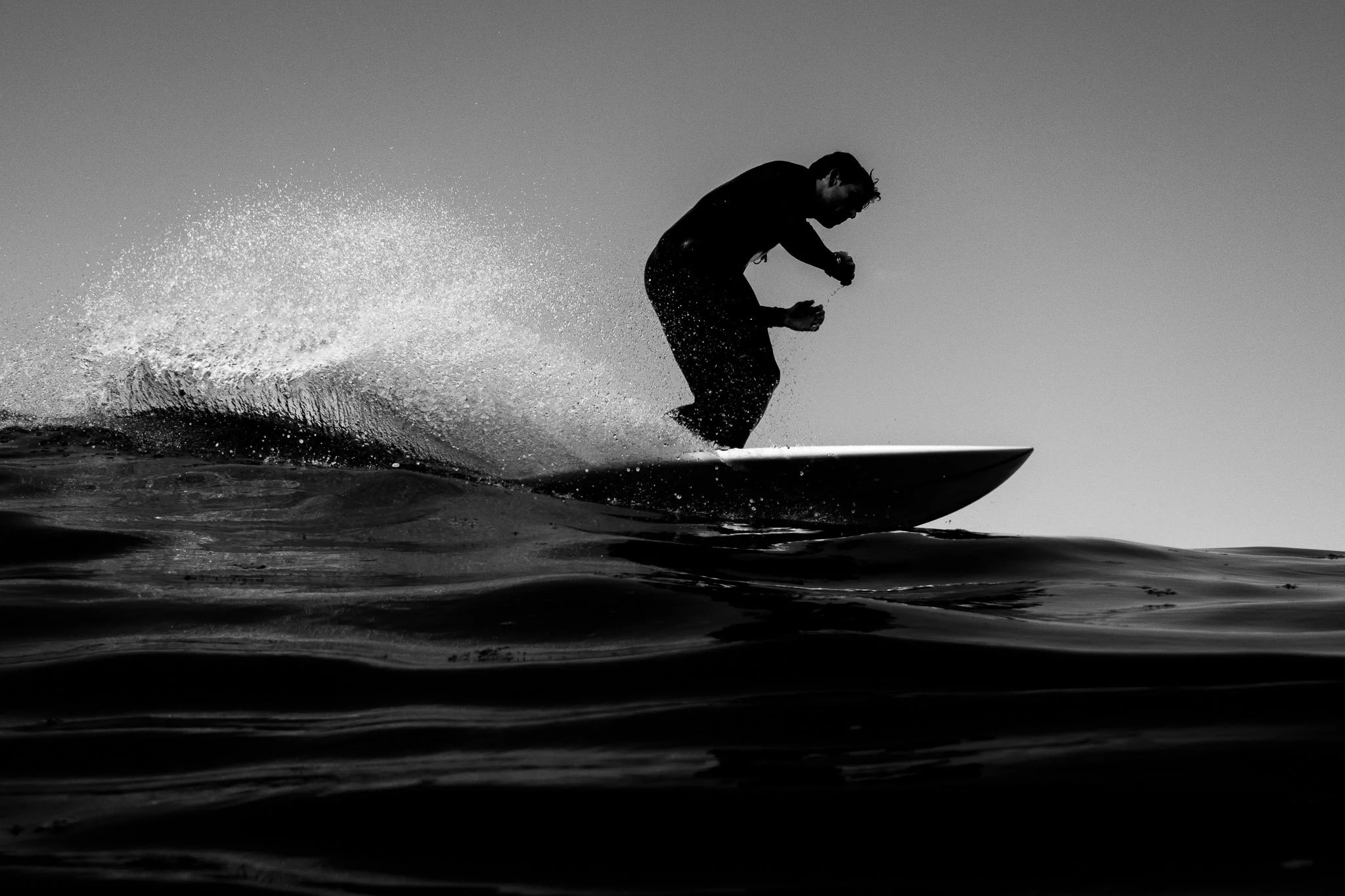 Silhouette of a person riding a surfboard on the water with a spray of water behind them, against a clear sky.