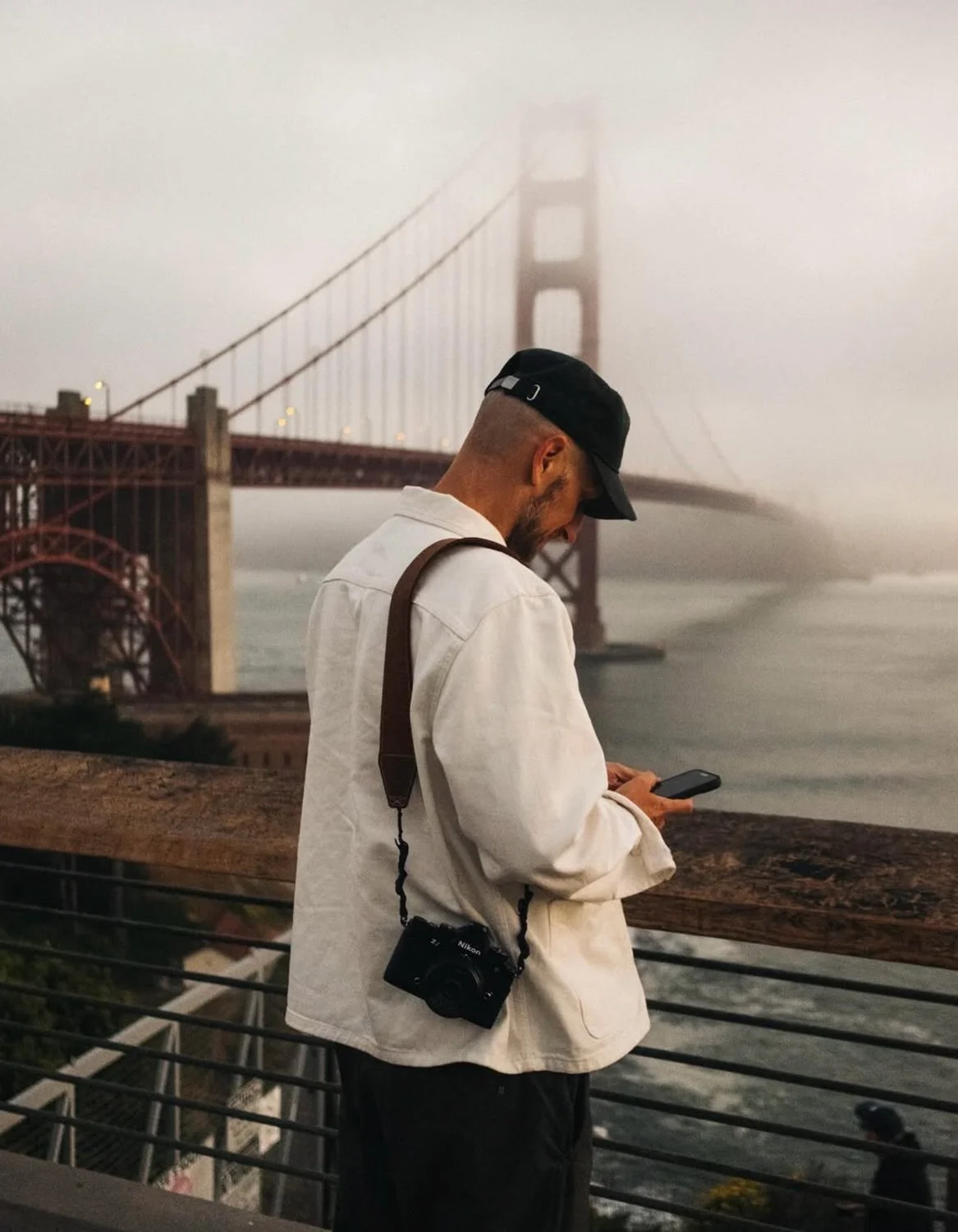 Nik Howe standing by a wooden railing looking at his phone with the Golden Gate Bridge shrouded in fog in the background.