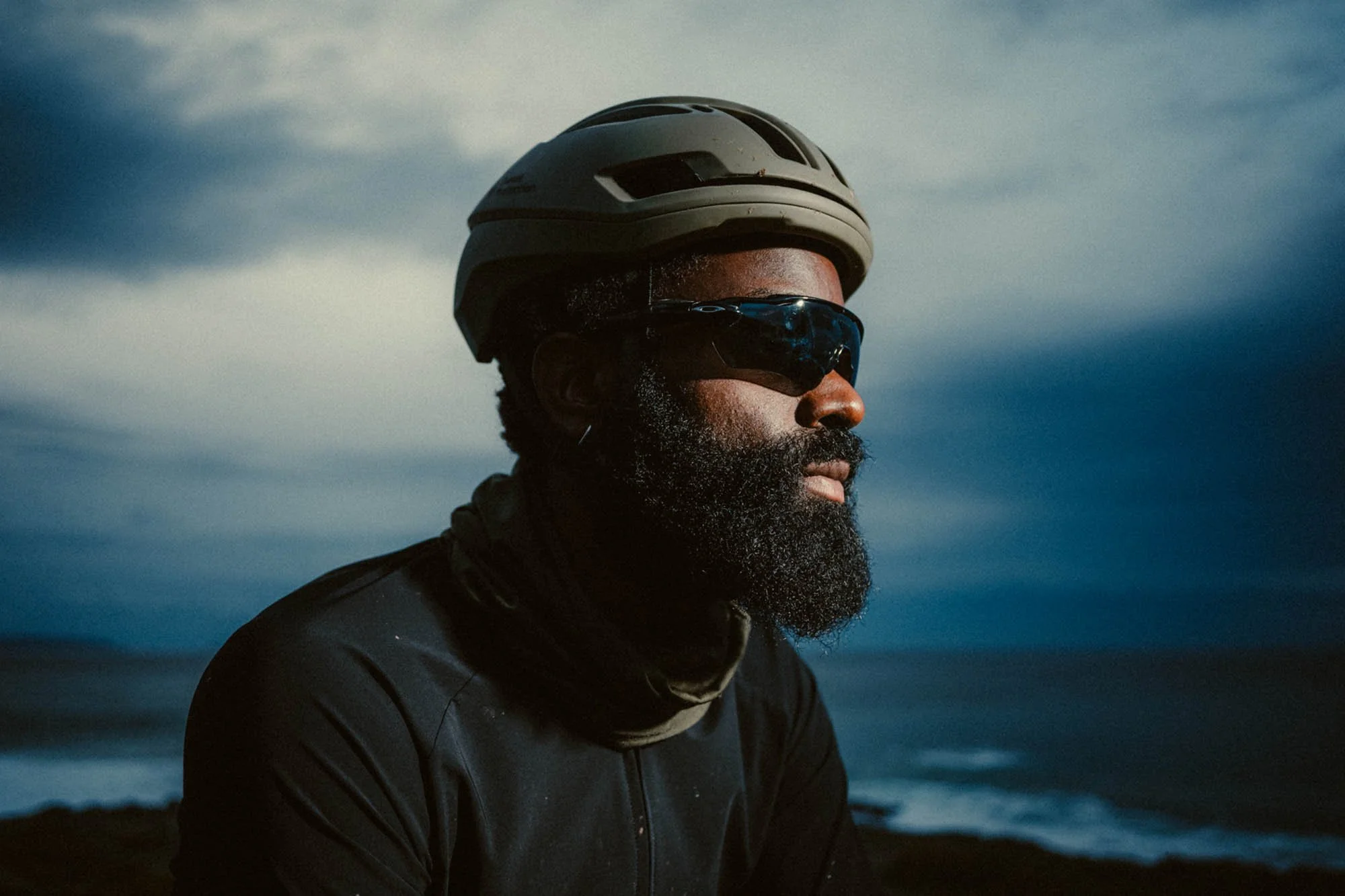 A man with a beard wearing a bike helmet and sunglasses outdoors at dusk or dawn, with a cloudy sky and dark landscape in the background. Shot for Zara Athleticz.