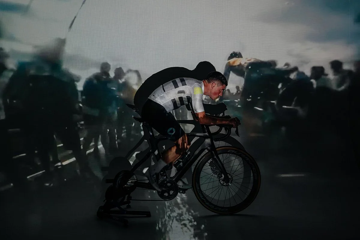 Pete Kennaugh riding a bicycle indoors with a large screen showing a crowd of people in the background.