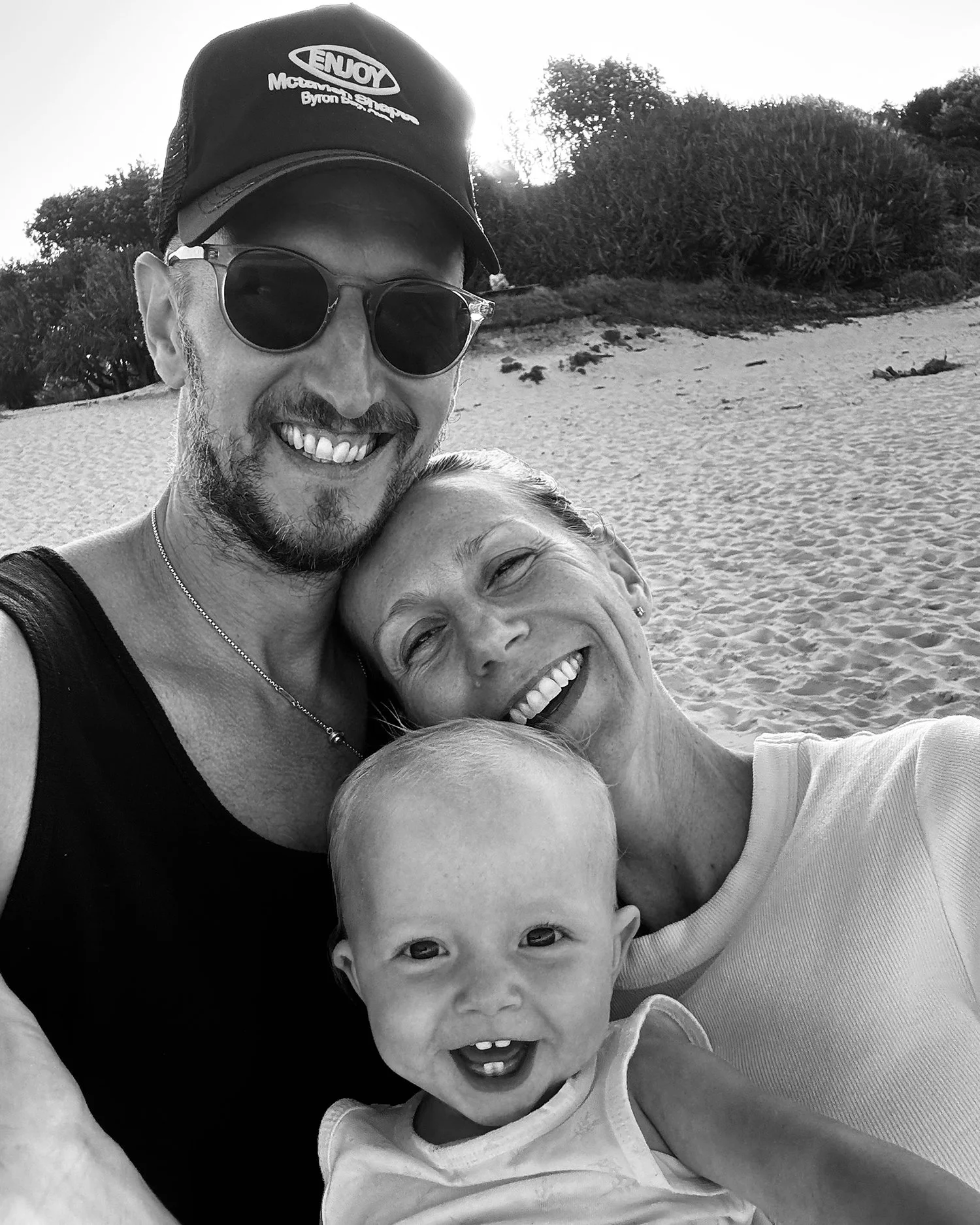 Nik Howe and family smiling and taking a selfie on a Byron Bay beach, with trees and bushes in the background, in black and white.