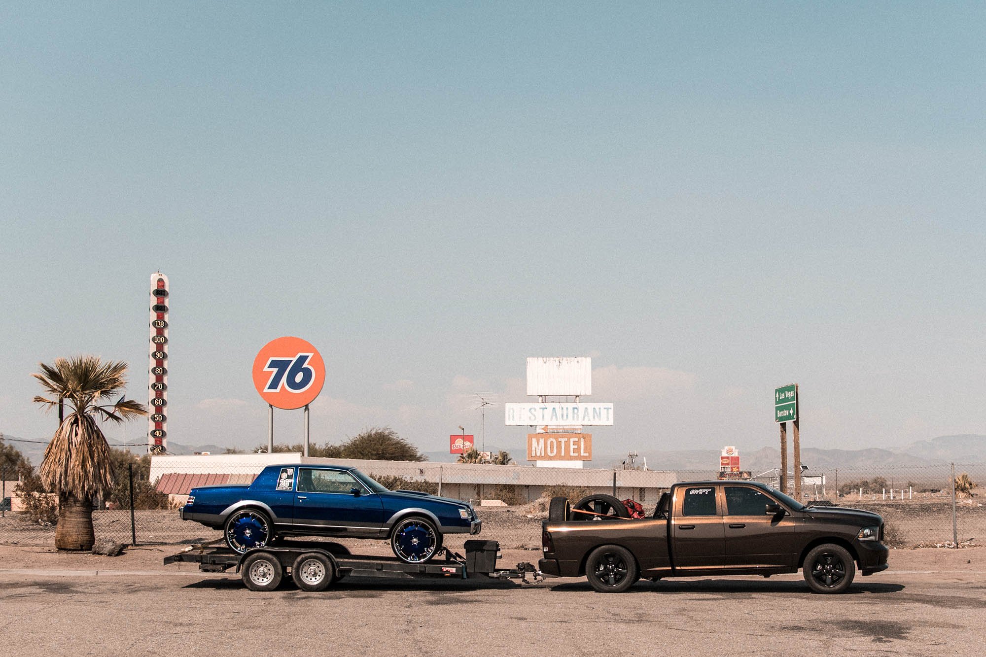 A black pickup truck towing a trailer with a blue classic car mounted on it. The background shows a desert landscape with a palm tree, a vintage gas station sign, and signs for 76 gas station and roads to Las Vegas and Barstow.