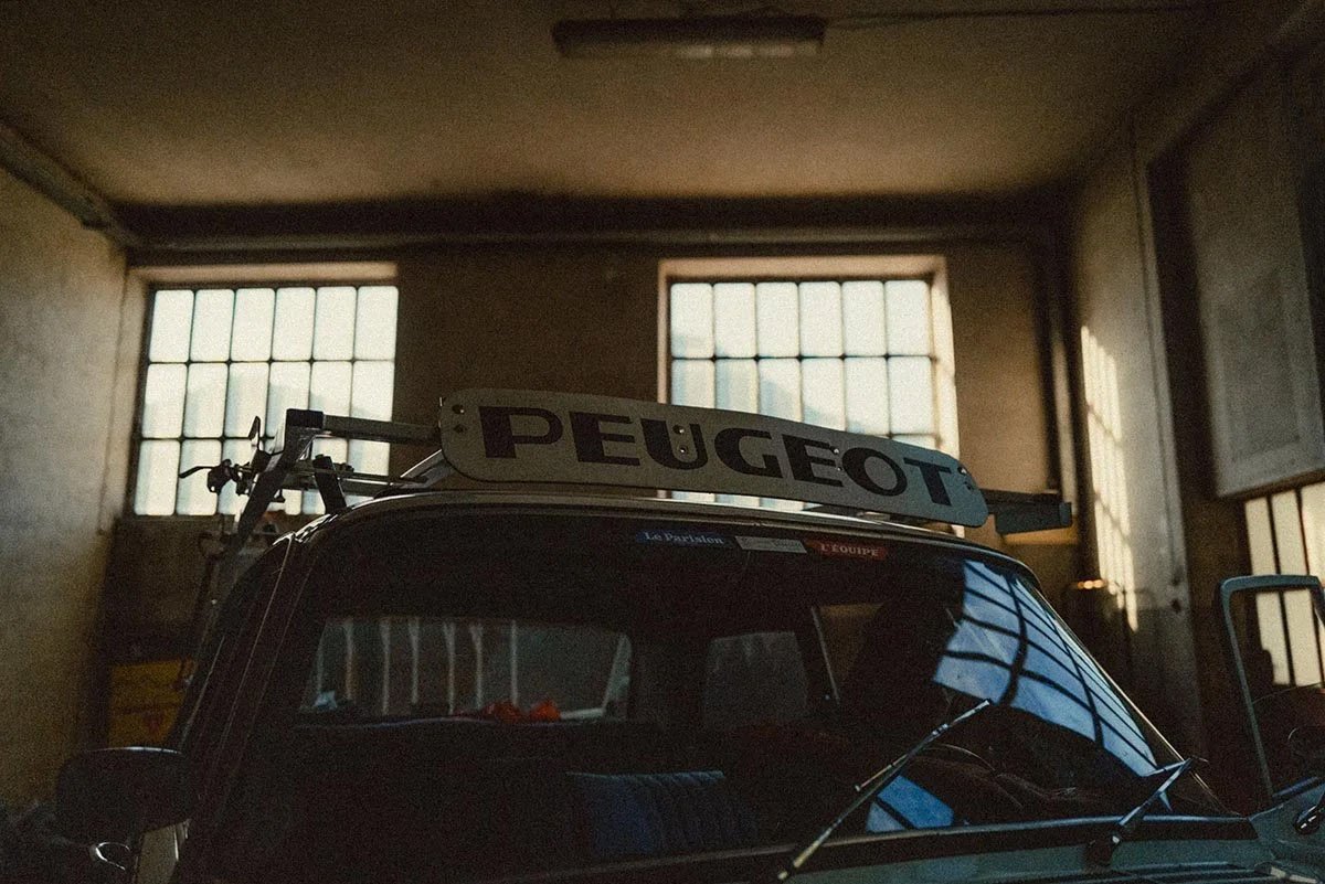 A vintage car inside a garage with a Peugeot sign on top, large multi-pane windows letting in natural light, and a bicycle mounted on the back of the car.