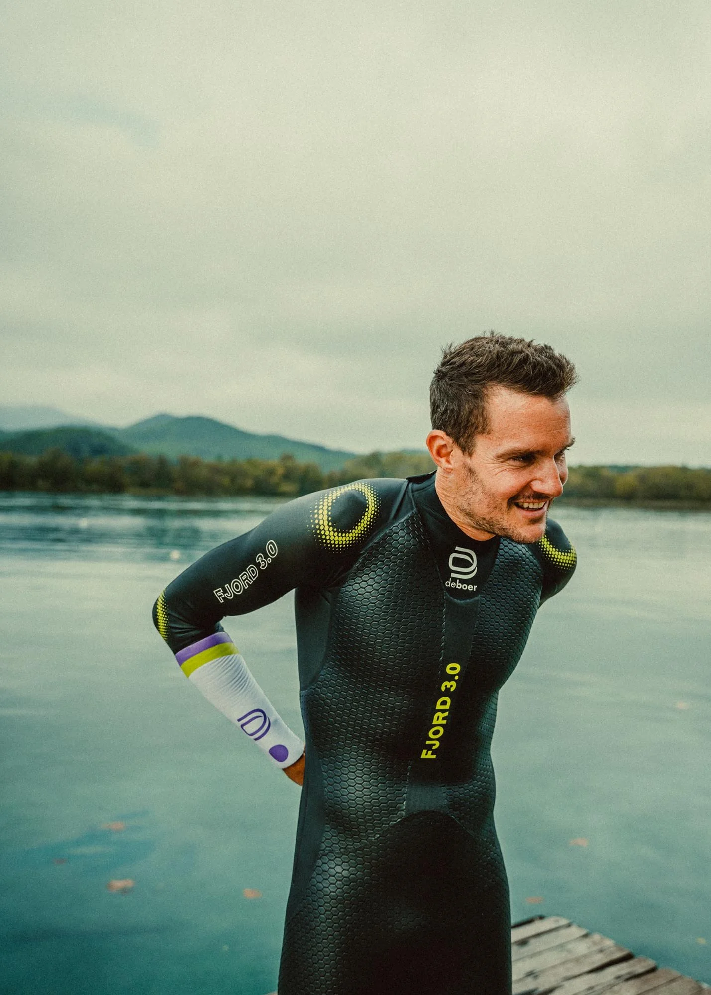 Jan Frodeno in a black DeBoer wetsuit with yellow and purple accents is standing on a dock by a lake, smiling and looking to the side, with mountains and cloudy sky in the background.