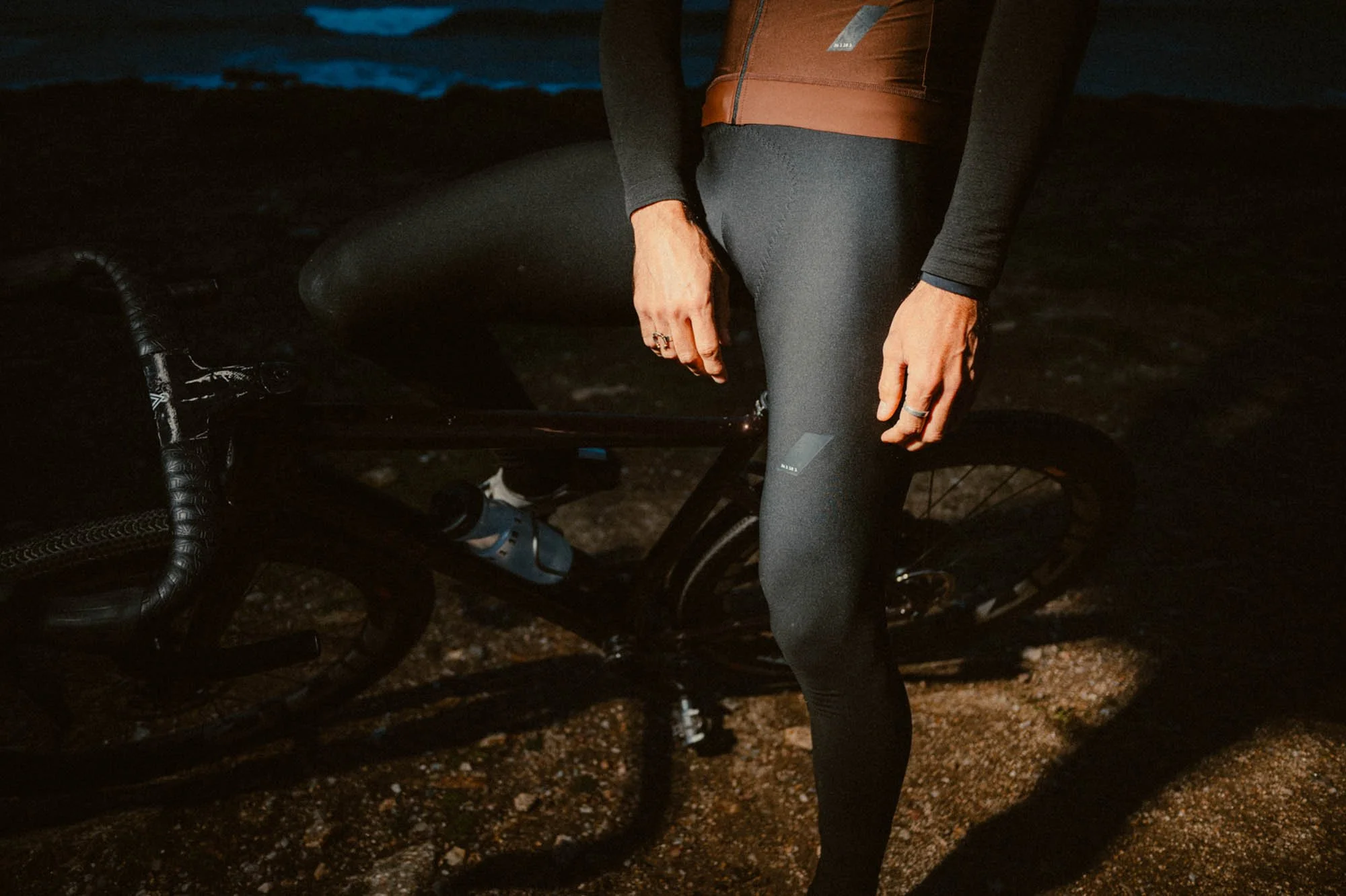 A man standing next to a bicycle on a gravel path at night, wearing black leggings and a brown and black jacket, with a water bottle attached to the bike. Shot for Zara Athleticz.