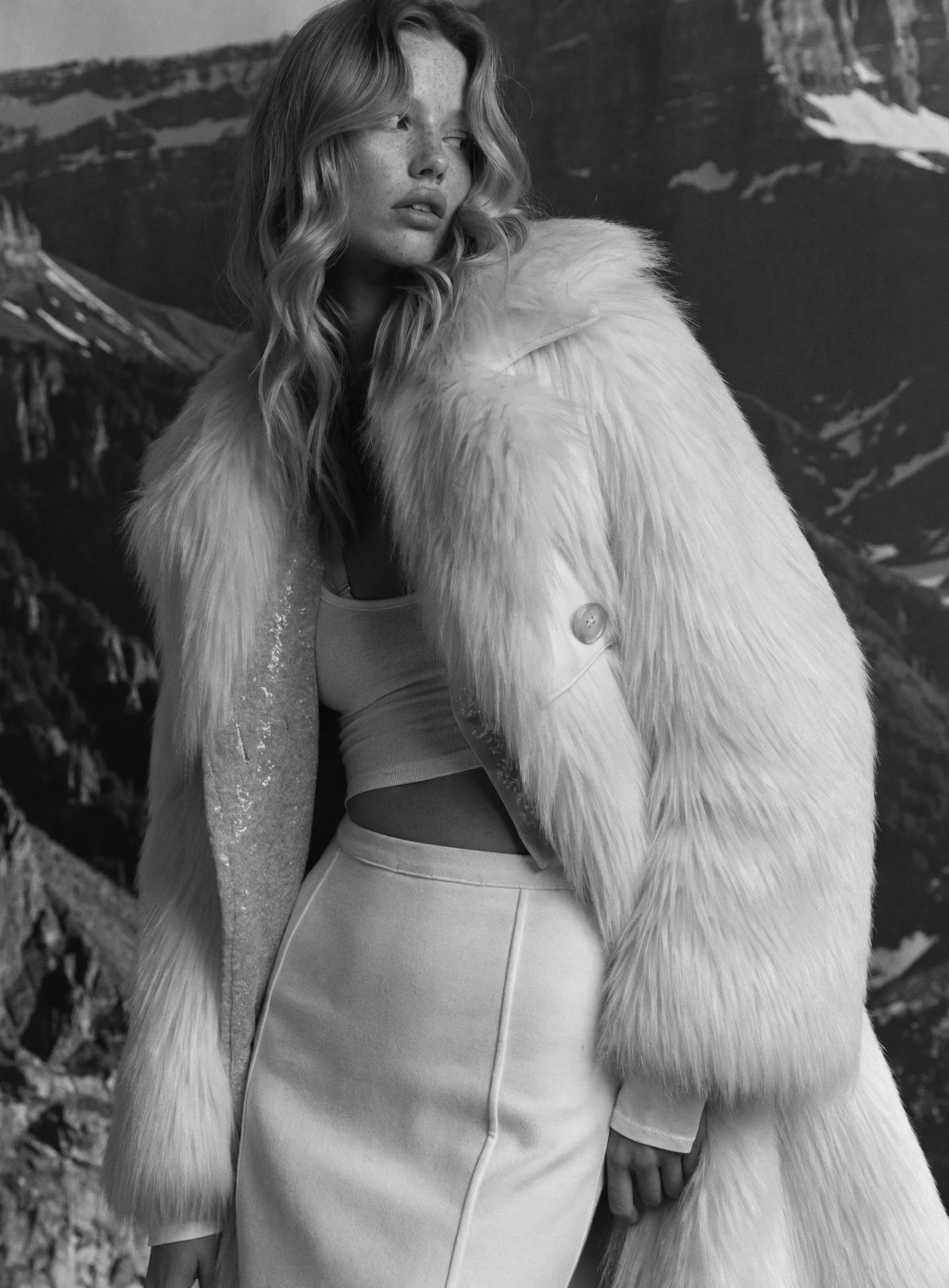 A woman with wavy blonde hair wearing a large fur coat over a tank top and skirt, standing in front of a mountainous landscape.