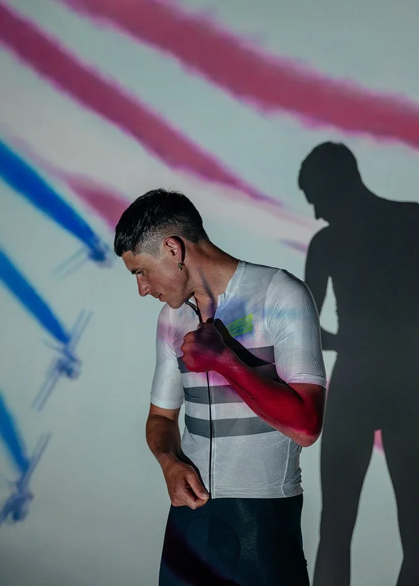 Pete Kennaugh adjusting his jersey in front of a wall with a colorful shadow pattern, with his shadow cast on the wall behind him.