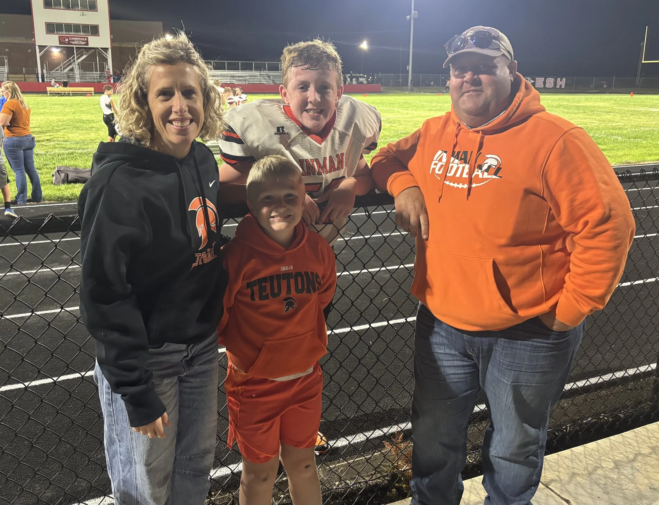 A group of four people, two adults and two children, at a football field during night. They are smiling, dressed in football and team apparel. The background shows a football field with players and a stadium with bright lights.