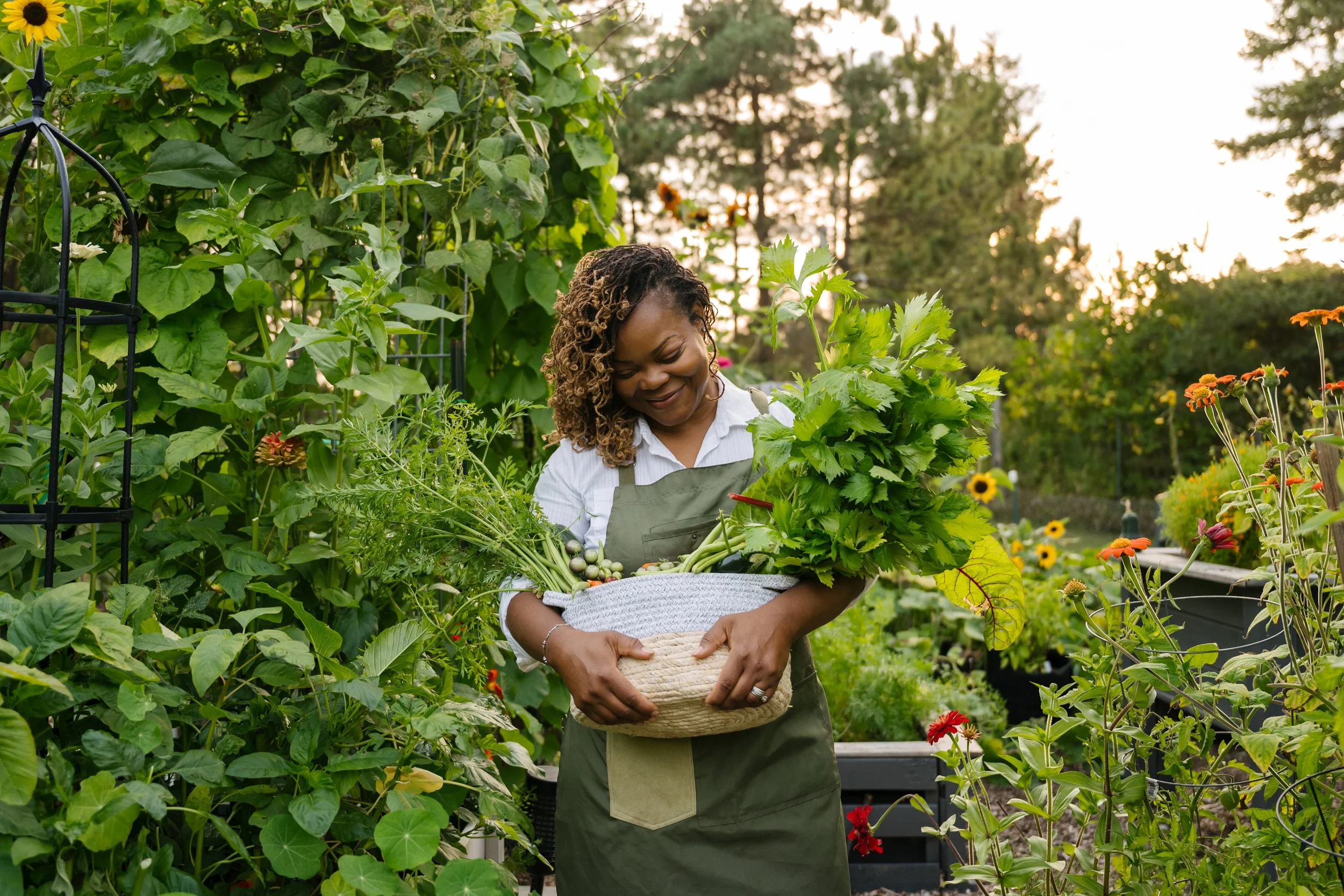 A woman in a garden holding a basket of freshly picked vegetables and herbs, surrounded by lush greenery and blooming flowers.