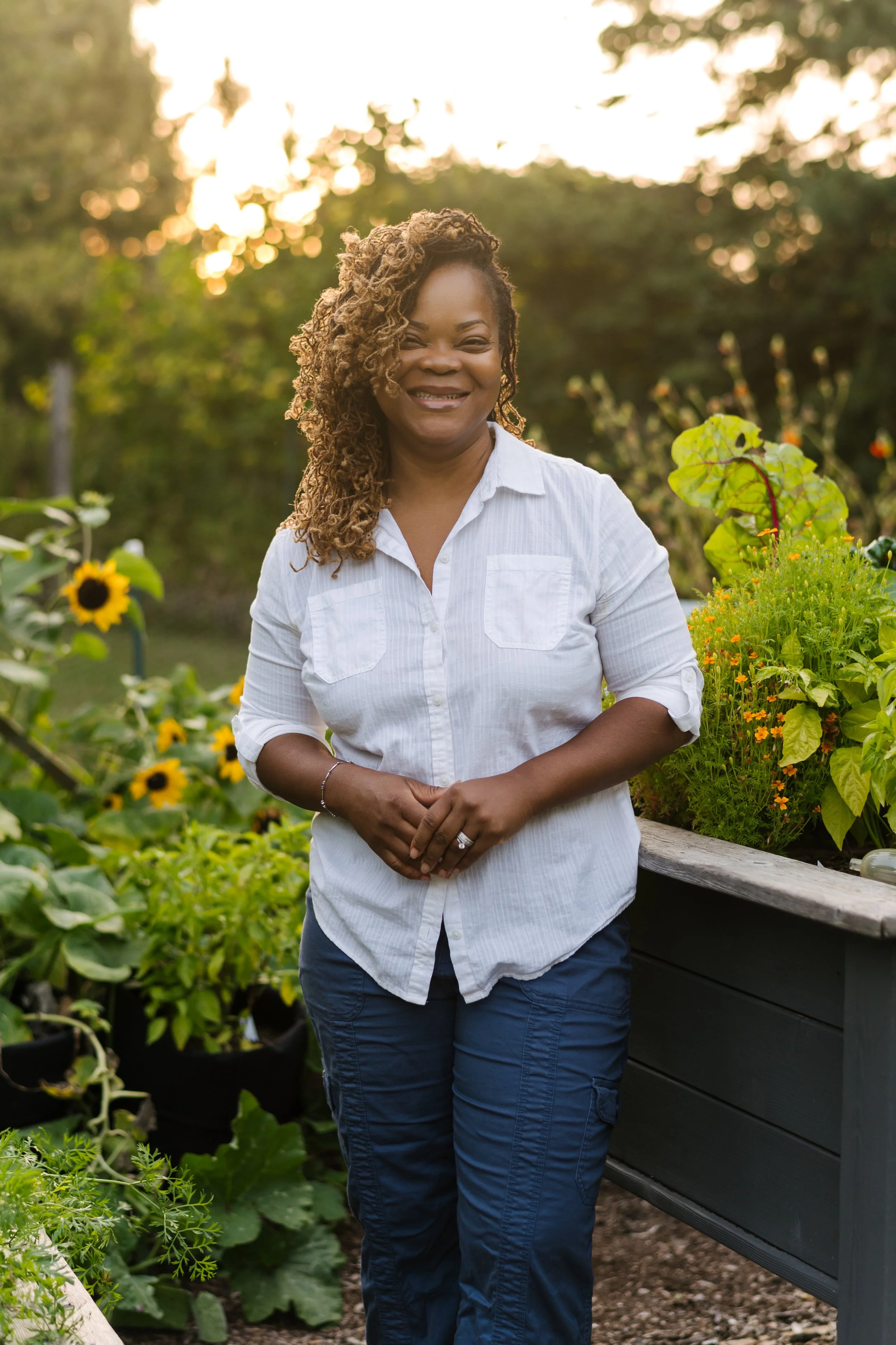A woman standing in a garden with sunflowers and other plants, smiling at the camera during sunset.