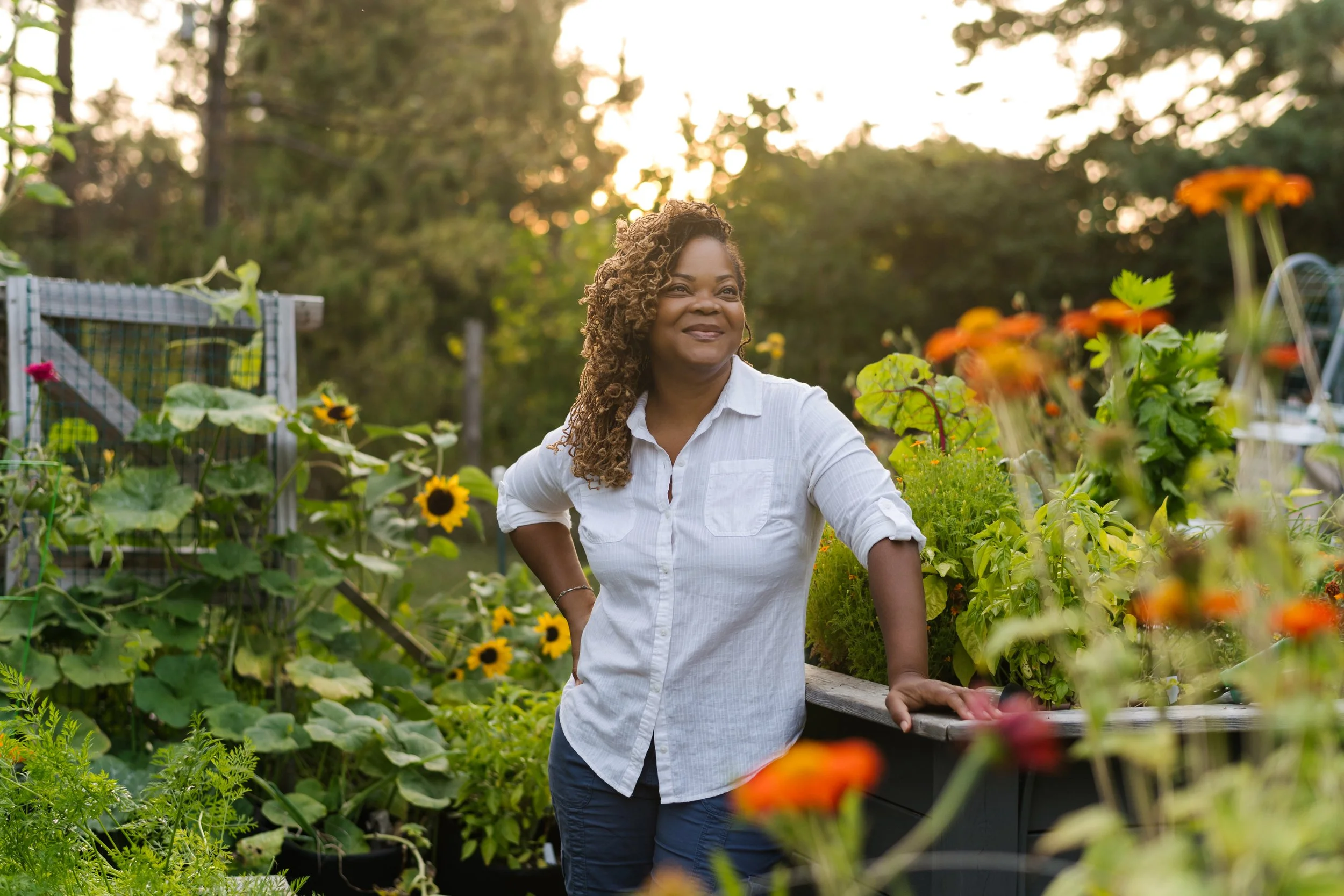 A woman with curly hair wearing a white shirt and blue jeans standing in a garden with flowering plants, sunflowers and greenery, with the sun setting in the background.