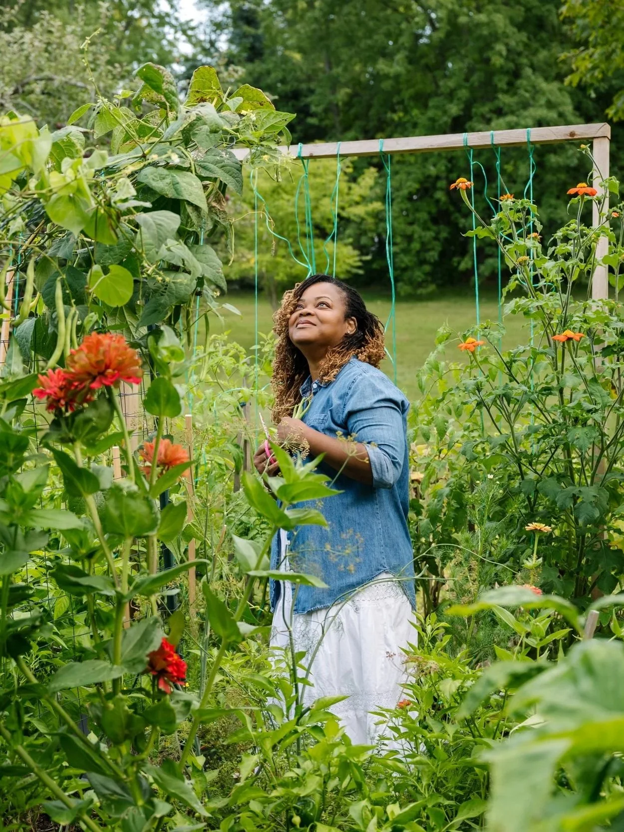 A woman in a denim jacket and white skirt standing in a lush green garden, smiling and holding a plant.