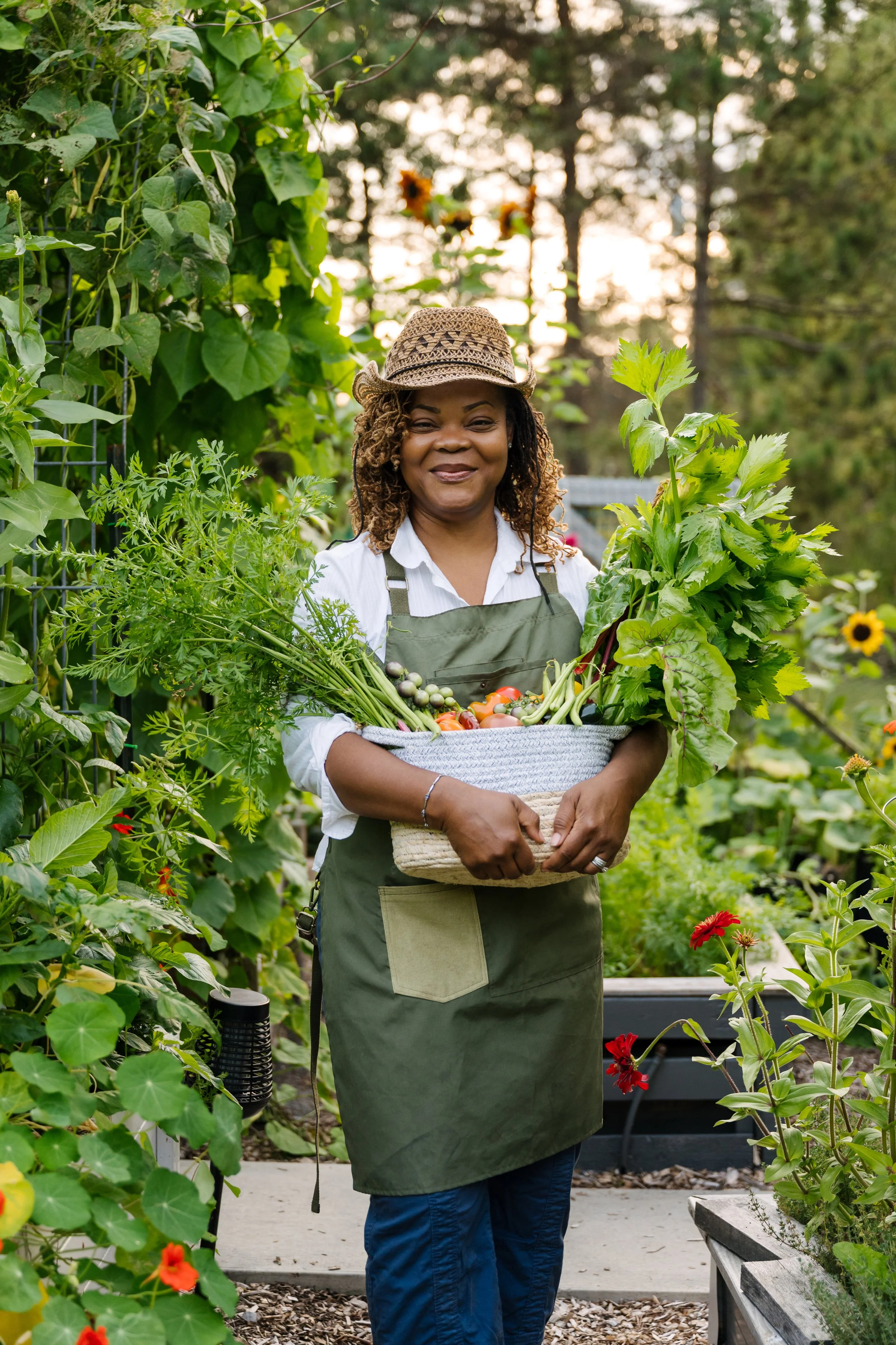 A woman standing in a garden holding a basket of freshly picked vegetables and herbs, smiling.