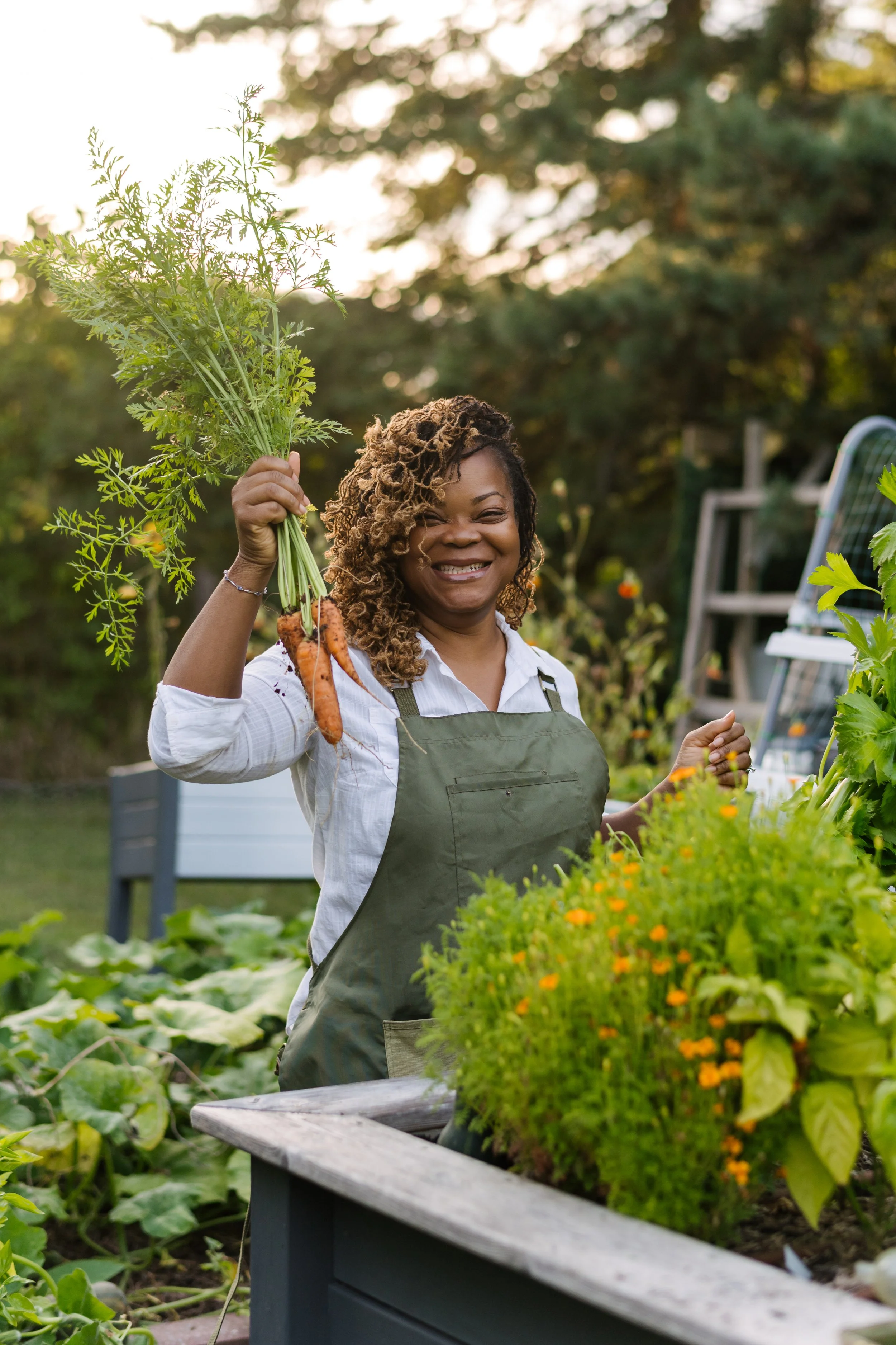 A woman smiling and holding freshly harvested carrots in a garden.