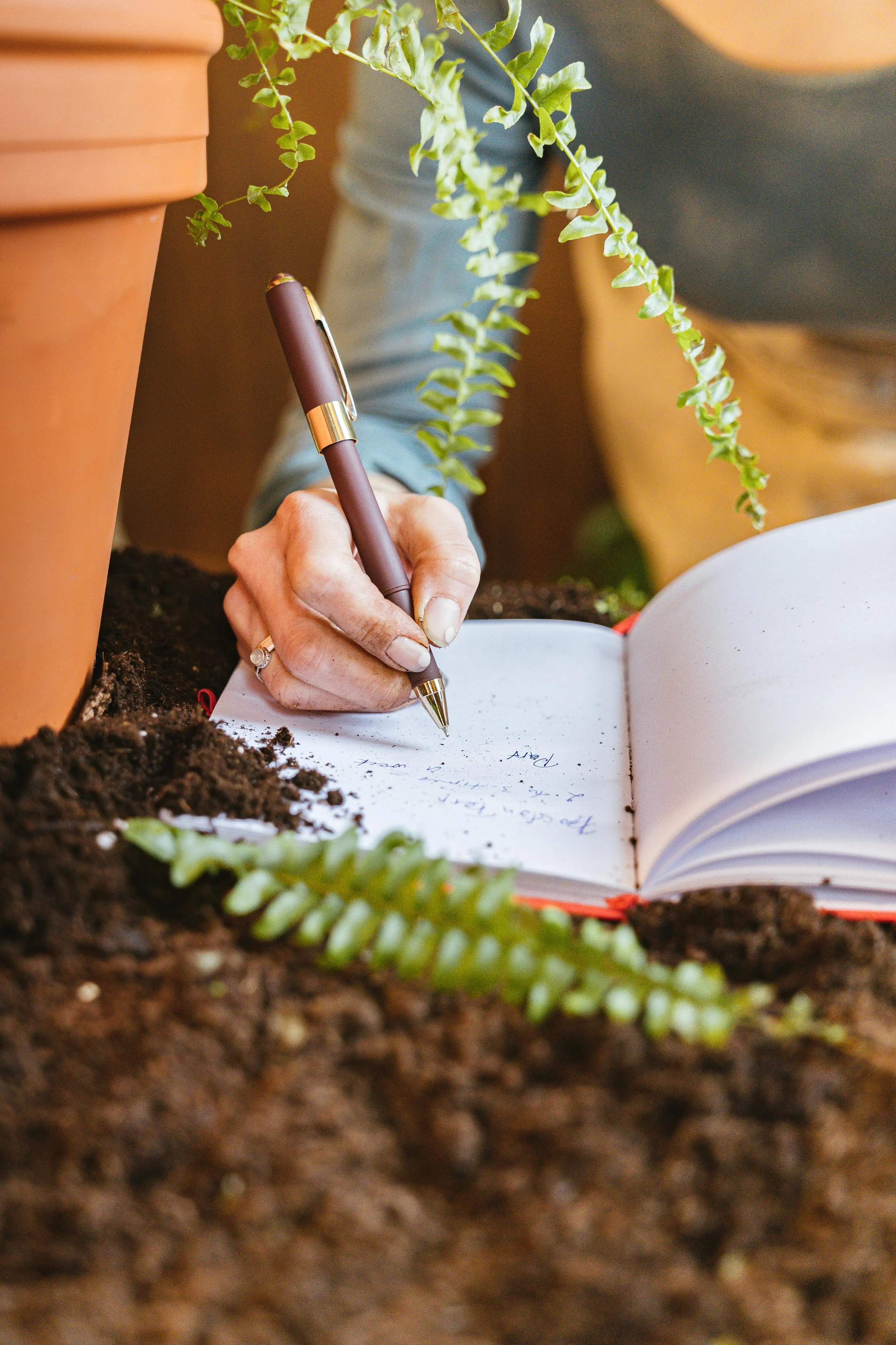 Close-up of a person writing in a notebook on soil, with potted plants and green fern in the background.