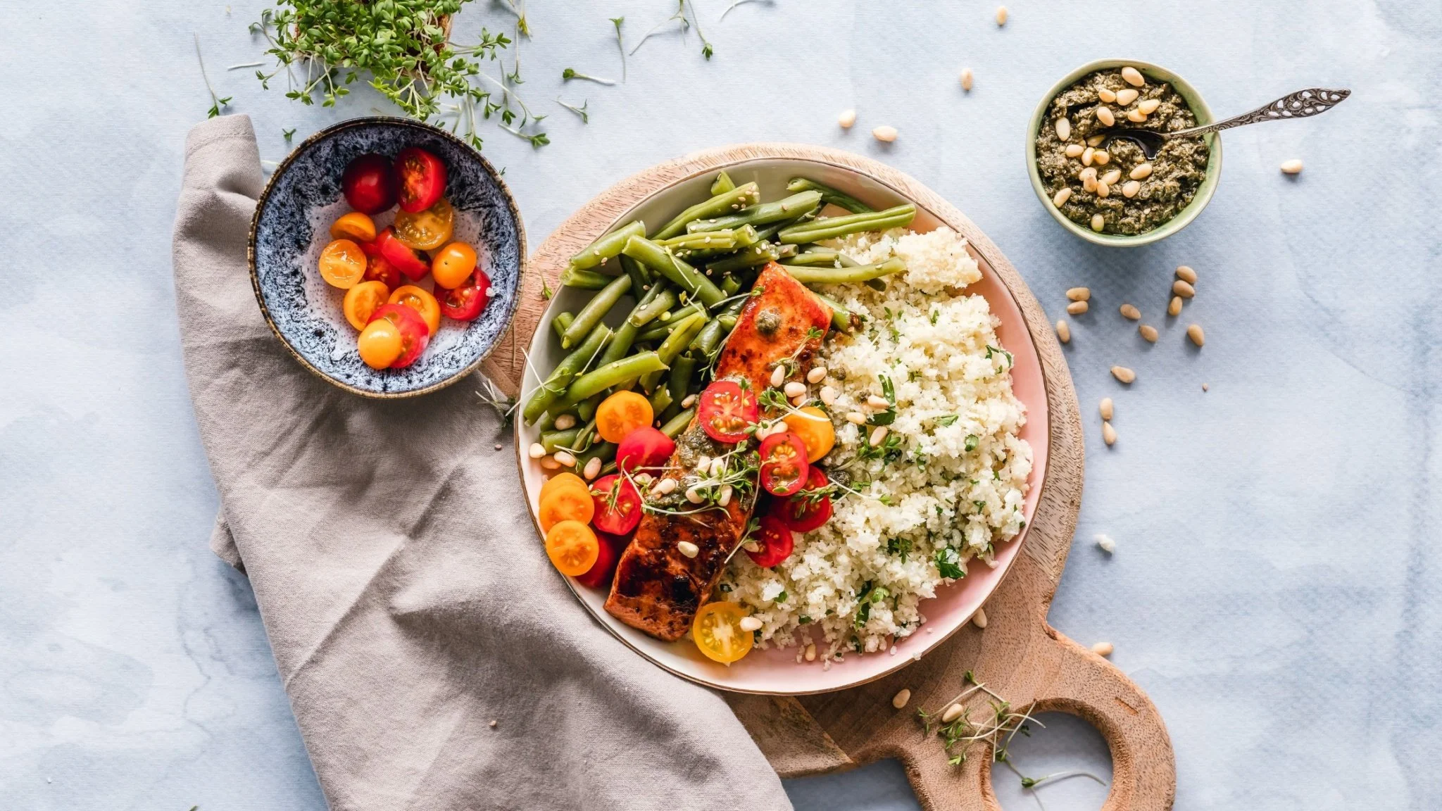 Plate with grilled salmon, green beans, cauliflower rice, cherry tomatoes, pine nuts, and herbs, with side bowls of cherry tomatoes and a pesto-like sauce, on a wooden cutting board.